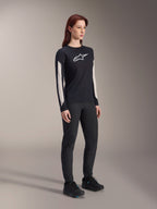 Woman wearing Alpinestars MTB gear, black Stella A-Dura Elite Jersey with white chest logo and sleeve panels, black Stella A-Dura Pants, black cycling shoes with blue accents, full performance outfit for mountain biking