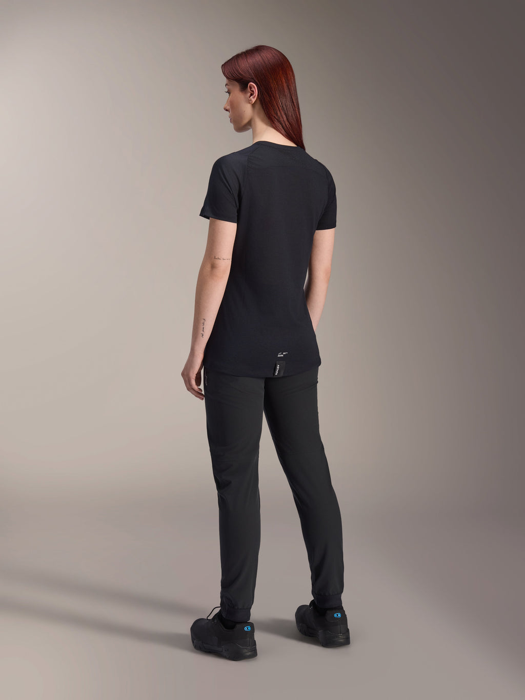 Woman model with red hair wearing the Alpinestars Stella A-Dura Dri Switch Short Sleeve Jersey in Black, rear view, paired with black mountain bike pants and shoes, showing the technical fit and longer drop tail for cycling coverage.
