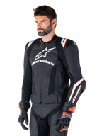 Male model wearing the Alpinestars Missile V3 Ignition Leather Jacket, Black with White and Orange accents, featuring prominent white logos, perforated leather sections, and protective shoulder armor, styled with racing gloves for a sport-racing look.