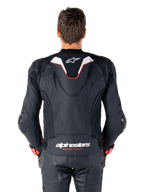 Rear view of a rider wearing the Alpinestars Missile V3 Ignition Leather Jacket, black leather with white and red accents, featuring a slim sport aero-hump with a white logo and 'alpinestars RACING AHEAD' branding on the lower back, paired with matching black leather racing pants and gloves.