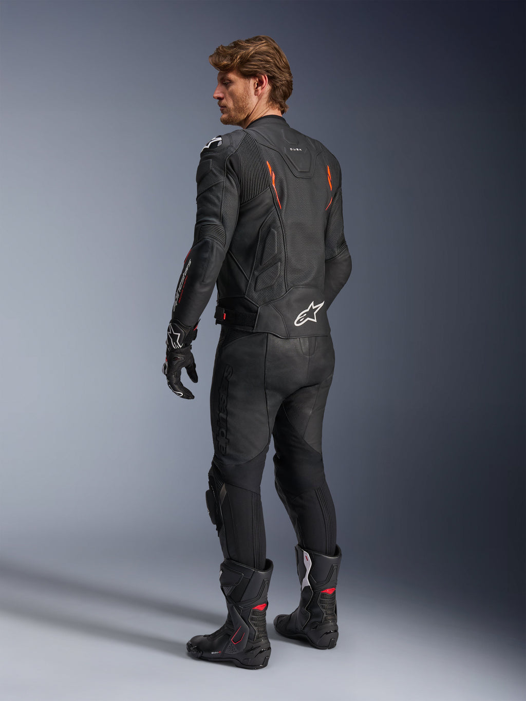 Alpinestars Dusk Airflow Leather Jacket, Black with Red Fluo accents, rear view of a rider in full leather motorcycle gear including matching black pants, gloves, and racing boots, featuring perforated leather and white Alpinestars logos.