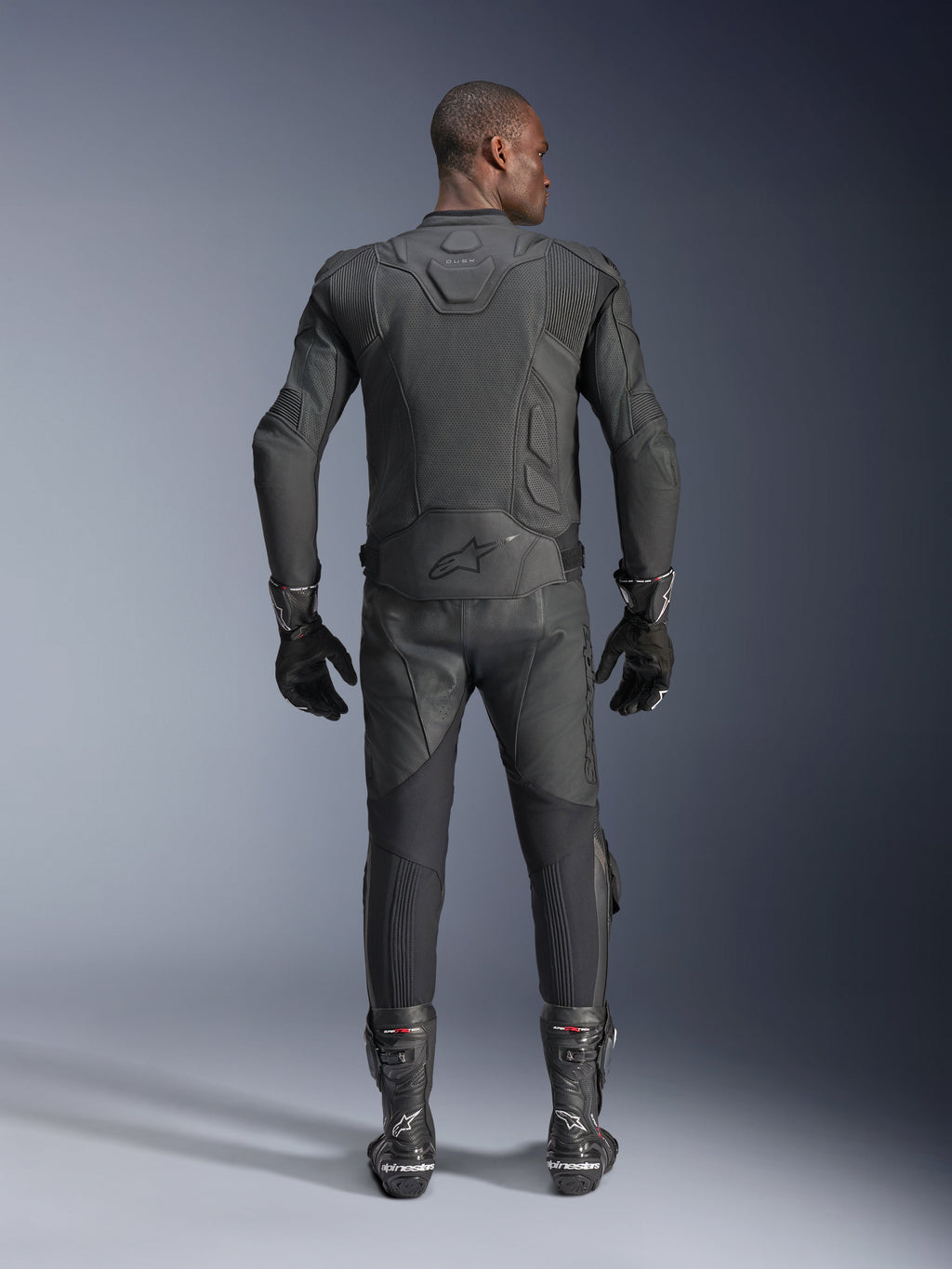 Male model shown from the back wearing the Alpinestars Dusk Airflow Leather Jacket in Black, paired with matching black leather racing pants, gloves, and boots, featuring extensive perforations for ventilation and the Alpinestars logo on the lower back.