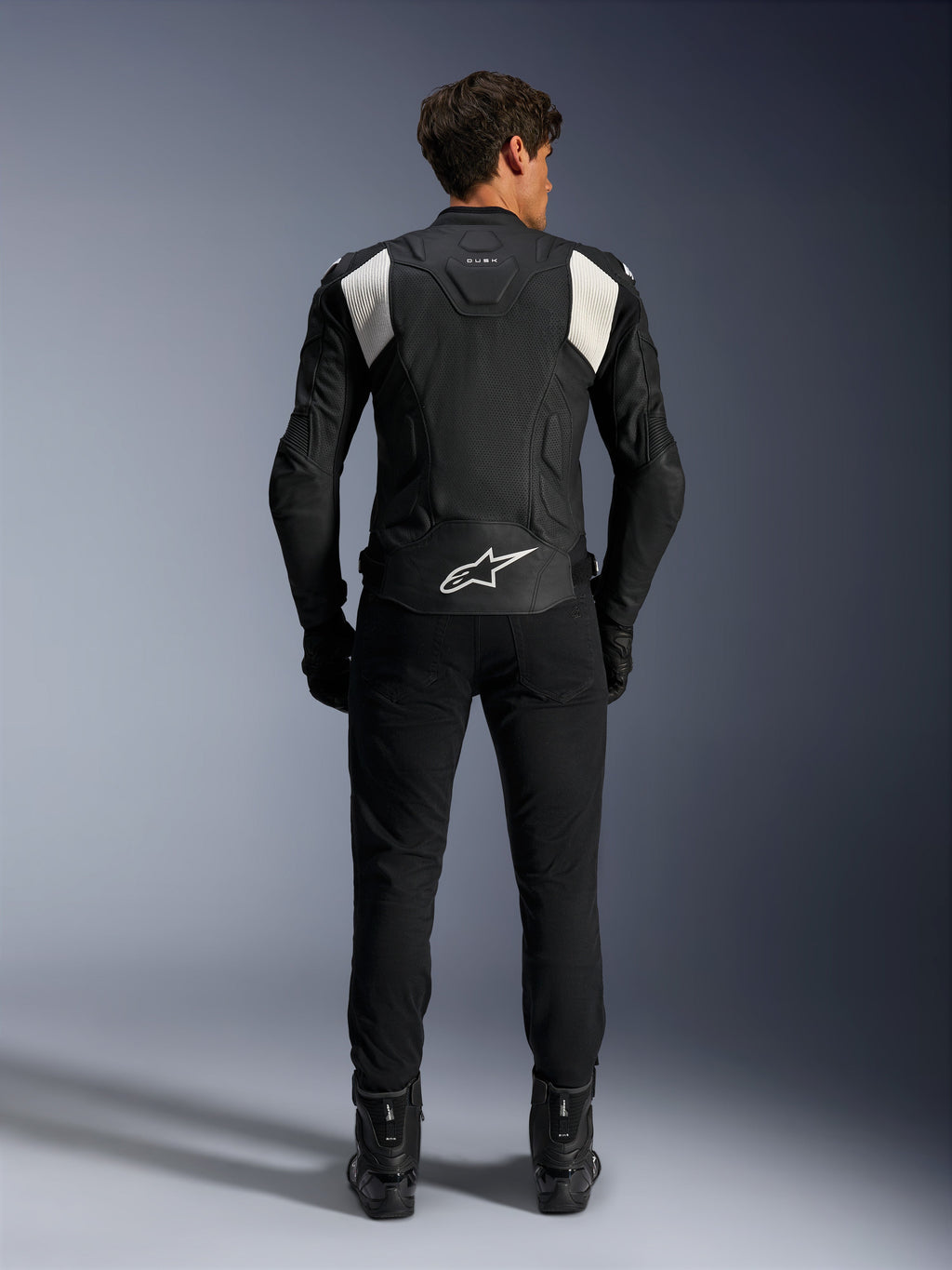 Alpinestars Dusk Airflow Leather Jacket, Black and White, rear view of a rider wearing black pants and boots, featuring perforated leather for ventilation, white accordion stretch panels on the shoulders, and white branding on the lower back