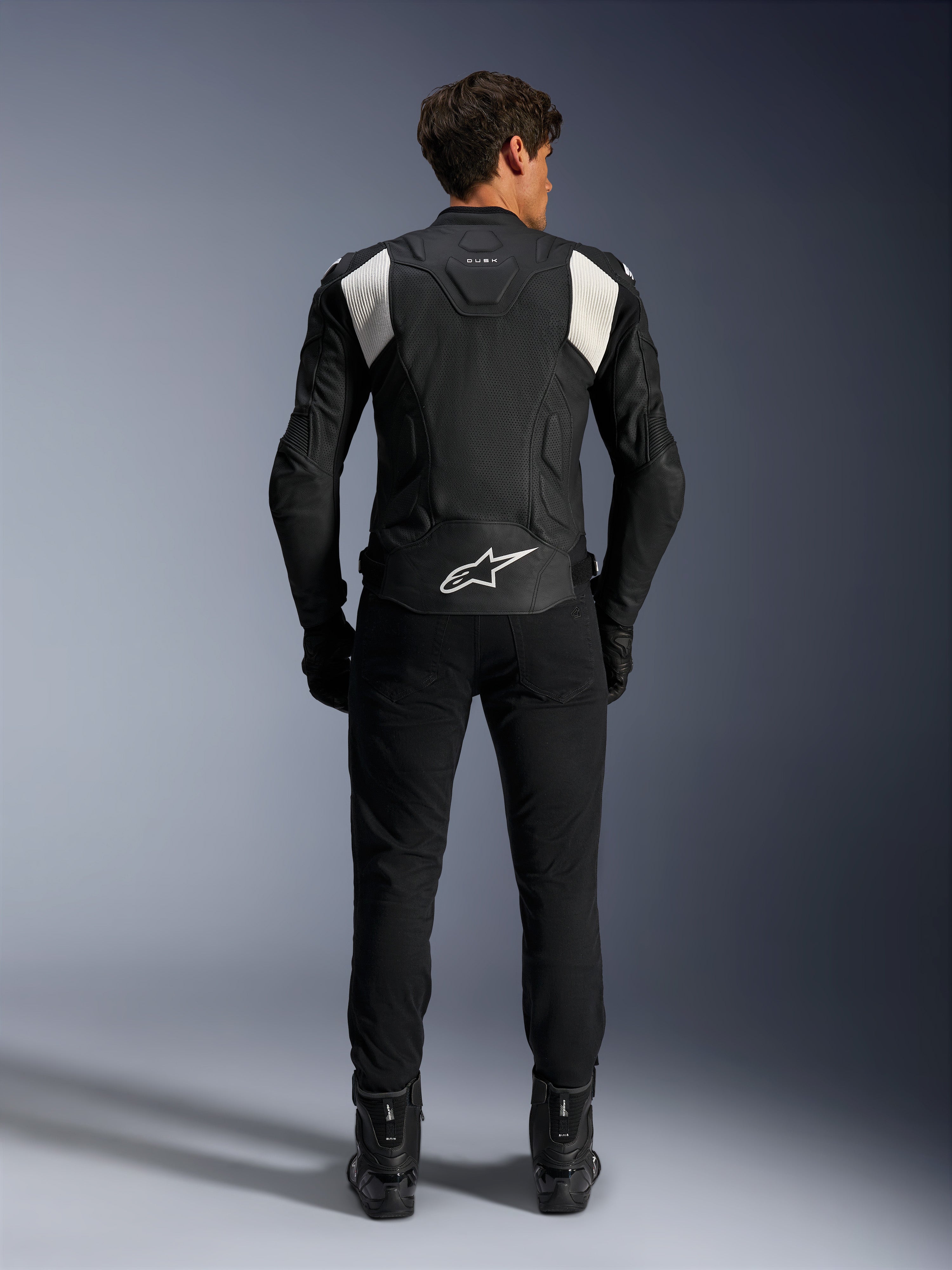 Alpinestars Dusk Airflow Leather Jacket, Black and White, rear view of a rider wearing black pants and boots, featuring perforated leather for ventilation, white accordion stretch panels on the shoulders, and white branding on the lower back