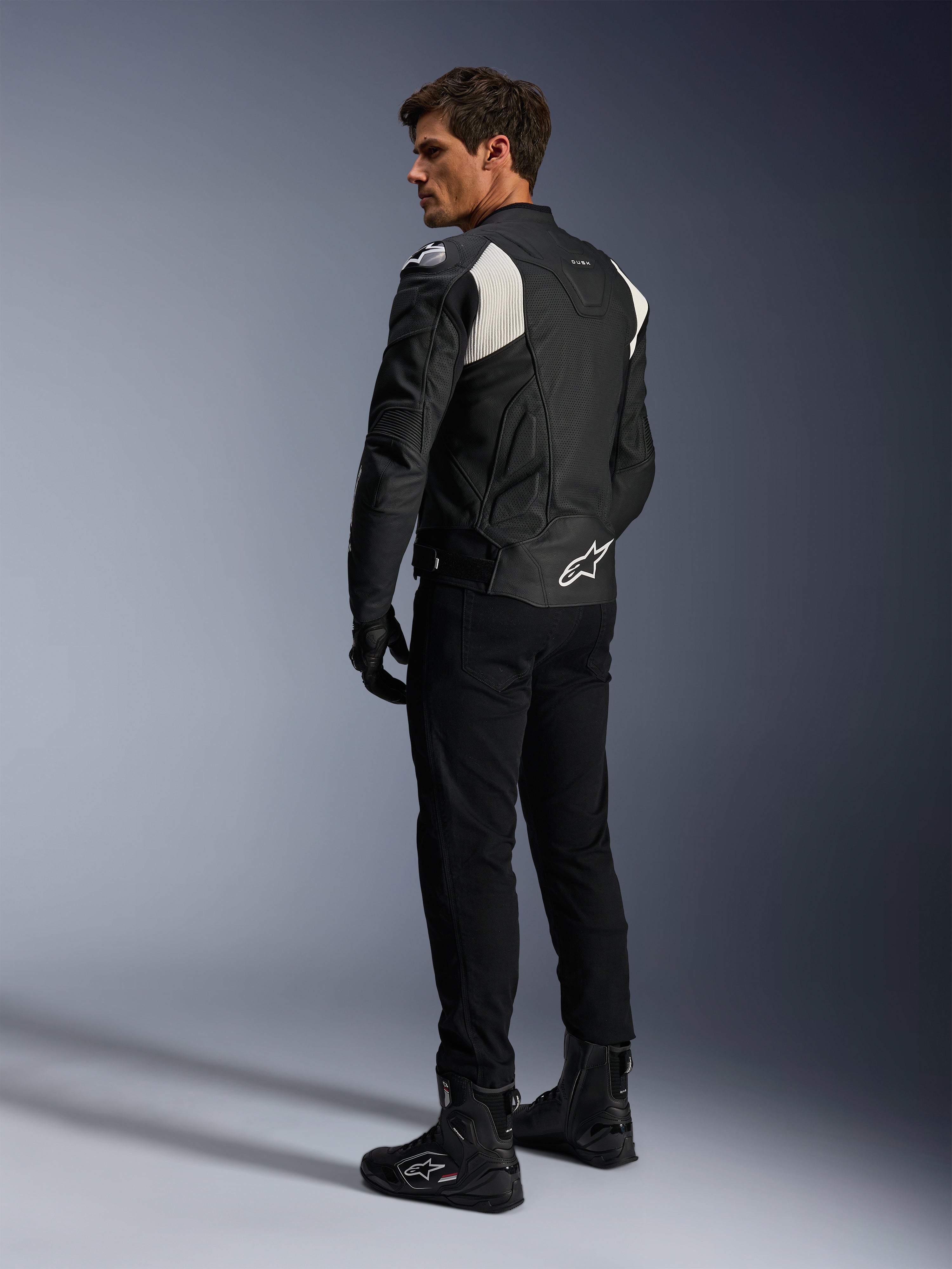 Alpinestars Dusk Airflow Leather Jacket, Black and White, rear view of model wearing the perforated leather jacket with matching black pants and boots, featuring white accordion stretch panels and bold logo on the lower back, sport motorcycle riding gear
