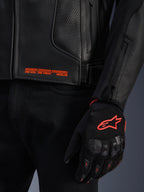 Alpinestars GP Plus R V4 Airflow Leather Jacket, close-up of black perforated leather with orange graphic text, paired with a black and orange motorcycle glove featuring the Astars logo