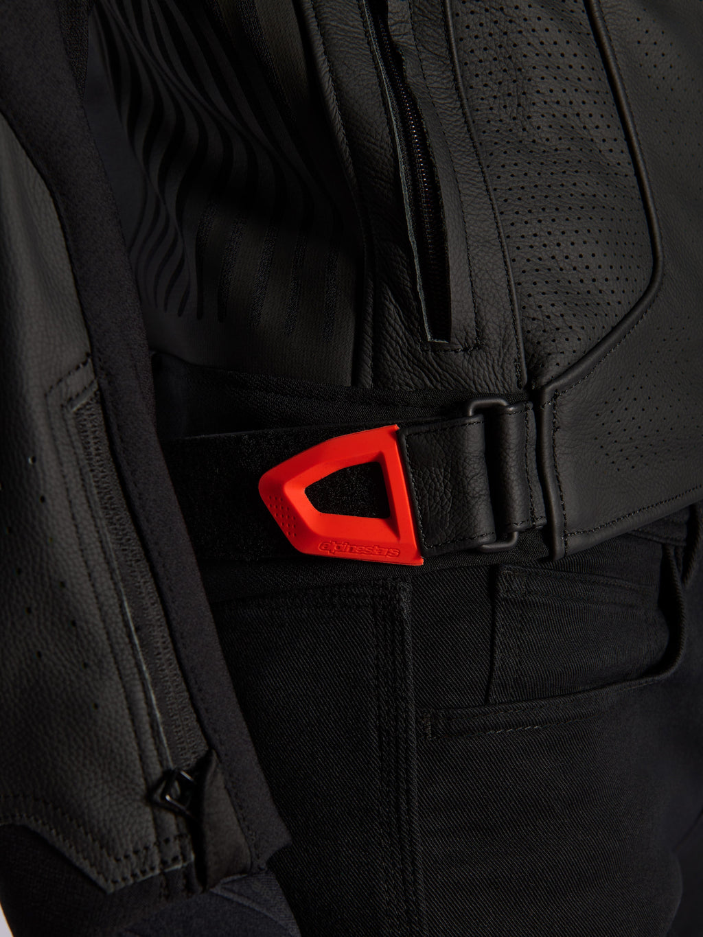 Alpinestars GP Plus R V4 Airflow Leather Jacket close-up, black perforated leather with a vibrant red fluo hook-and-loop waist adjustment strap, featuring a zippered side pocket and ergonomic textured panels.