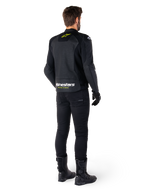 Rear view of a rider wearing the Alpinestars Faster V3 Airflow Leather Jacket, Black with White and Yellow Fluo accents, paired with black pants and boots, featuring perforated leather for ventilation and bold 'RACING AHEAD' branding on the lower back.
