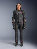 A woman wearing the Alpinestars Stella Dusk Leather Jacket in Black Diva Pink White, featuring a white chest logo and pink accents on the shoulders and sleeves, paired with black motorcycle pants, gloves, and boots.