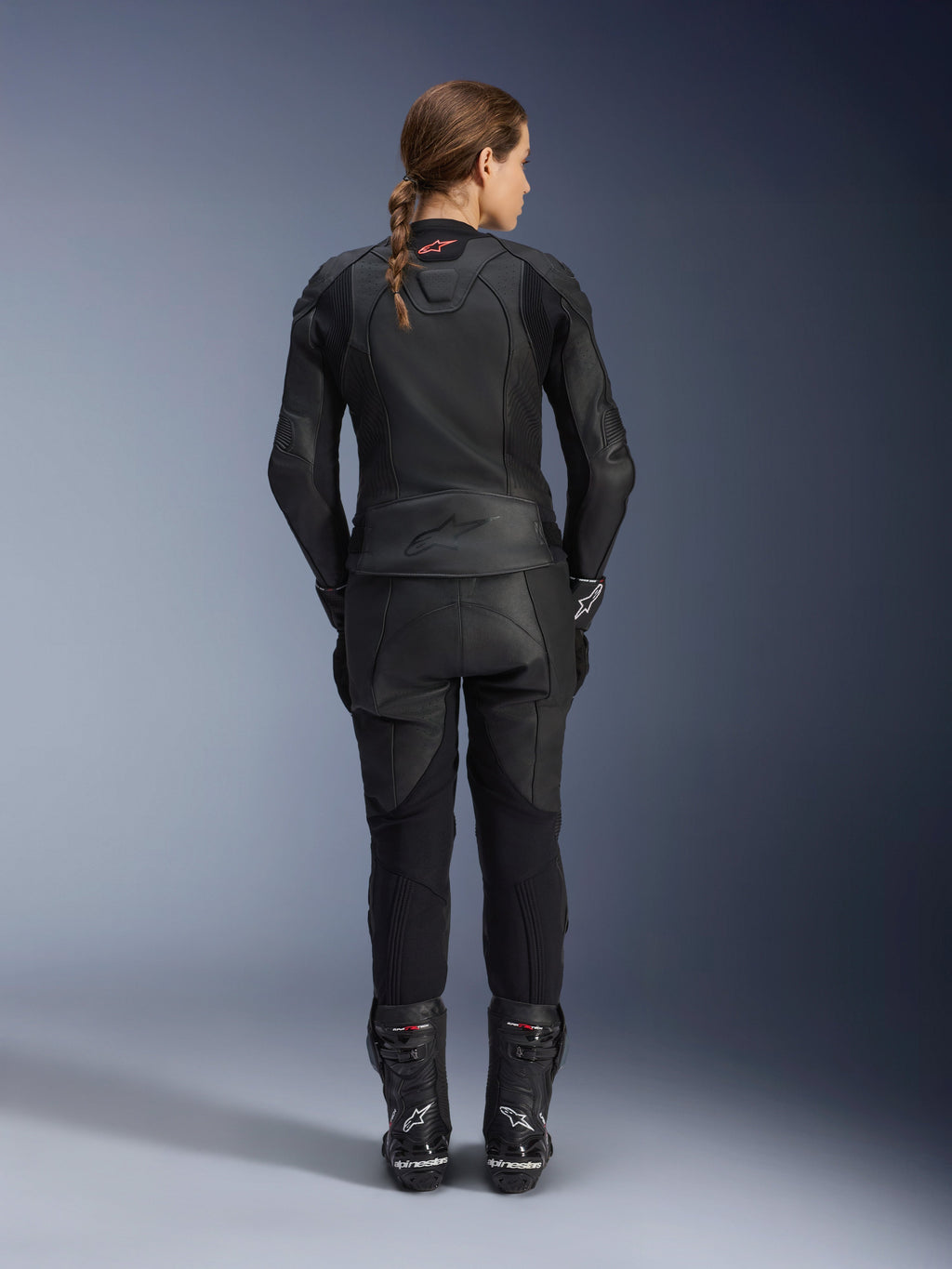 Alpinestars Women Stella GP Plus V4 Leather Jacket, Black Black, full-body rear view of a female rider wearing matching leather pants and boots, sleek minimalist design with a small red logo on the neck, tailored sport silhouette for women