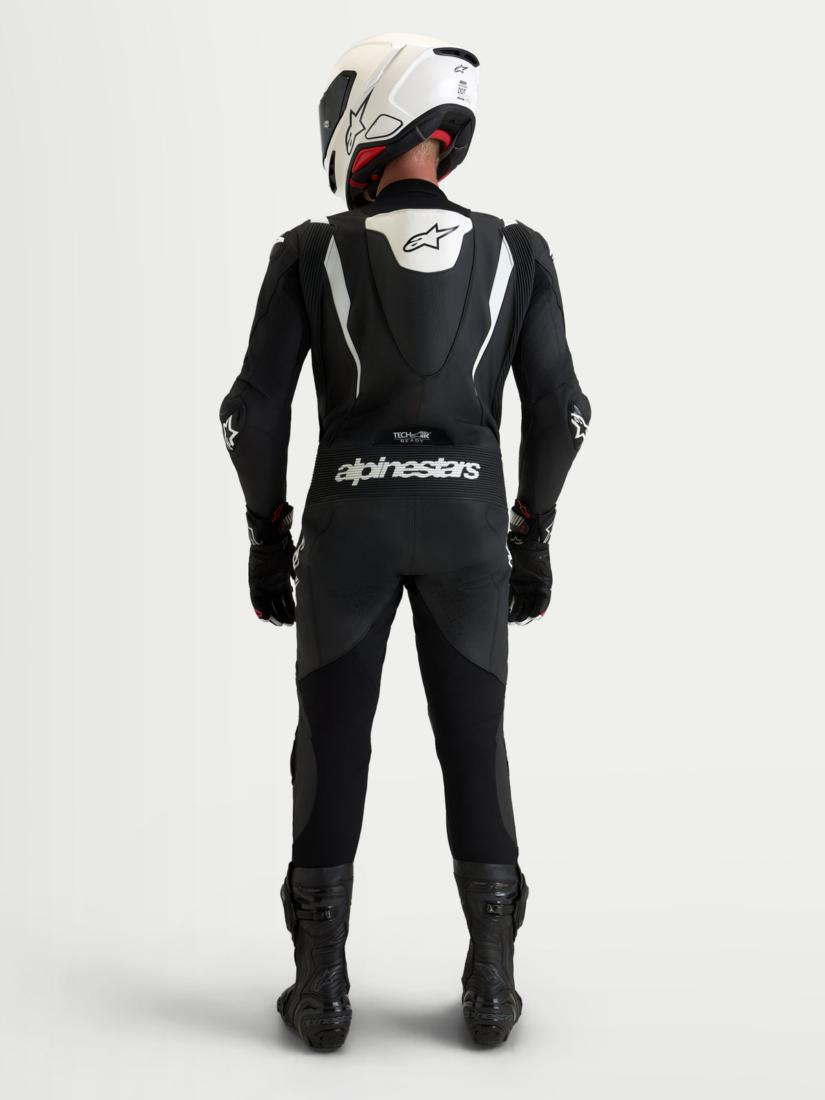 Alpinestars Missile V3 Leather Suit 1 Pc, Men's 1-piece motorcycle racing suit, Black and White, featuring an aerodynamic speed hump, CE AAA-rated leather construction, and Alpinestars branding across the back, shown from behind on a rider wearing a white helmet and black boots.
