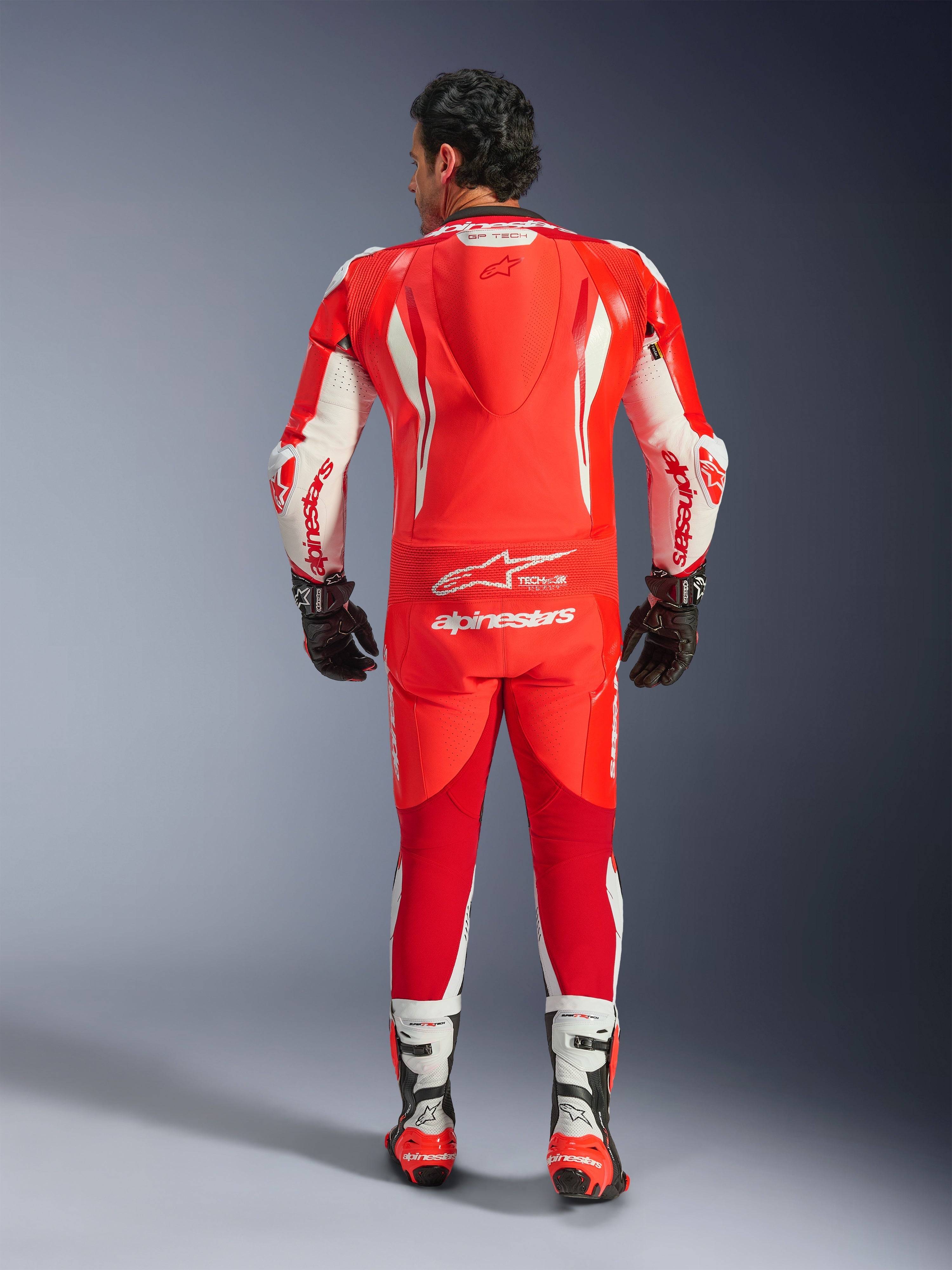 GP Tech V5 One Piece Leather Suit, motorcycle racing suit, Red Fluo Bright Red and White, back view showing aerodynamic hump and white Alpinestars branding, worn with black and white racing gloves and boots.