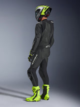 Motorcycle rider wearing Alpinestars GP Force V2 Leather Suit 1-Piece in Black, viewed from the back-side with white branding on the thigh and lower back, aerodynamic speed hump, paired with a neon yellow and black helmet, matching racing gloves and boots, professional track and sport riding gear.
