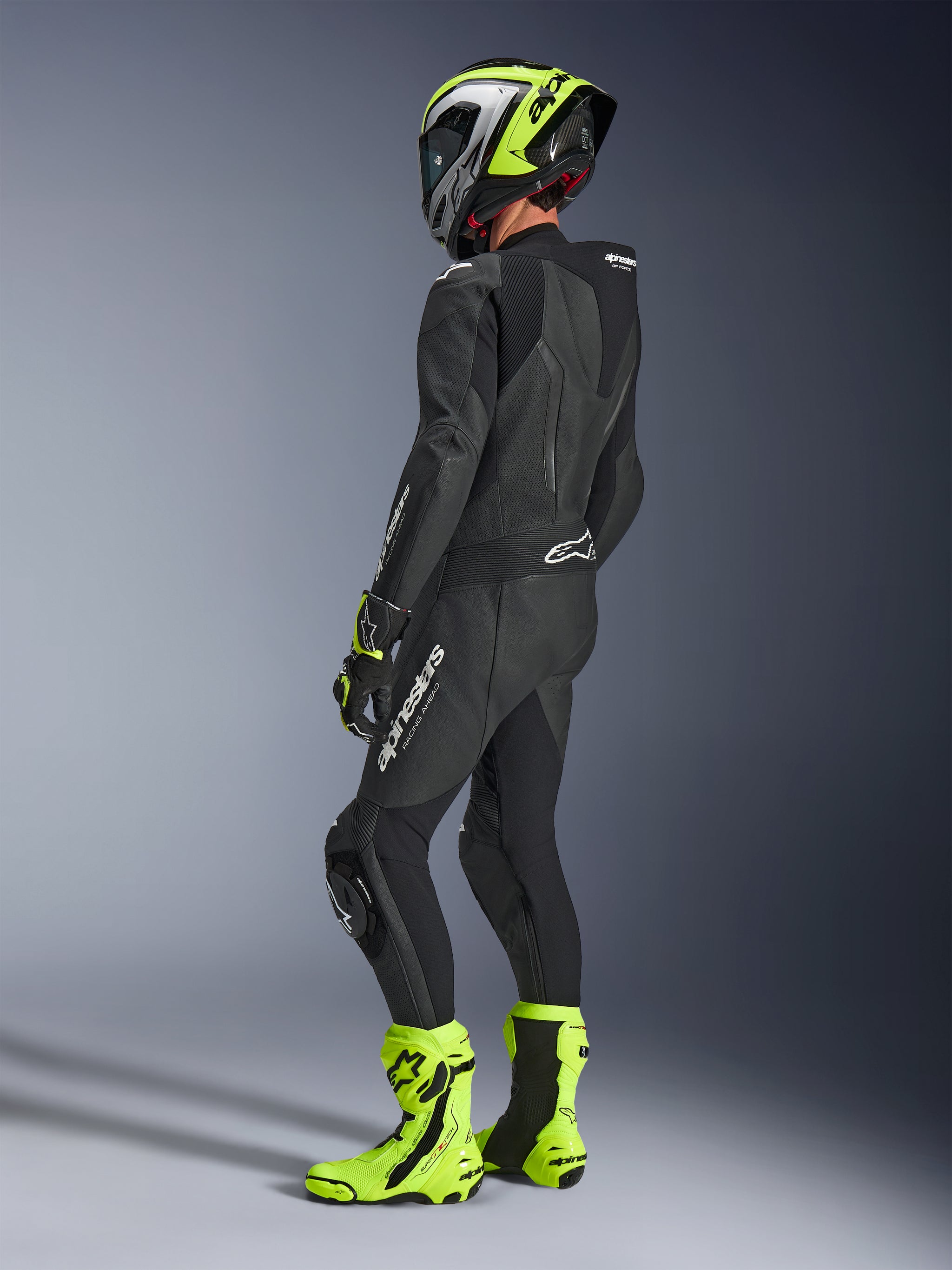 Motorcycle rider wearing Alpinestars GP Force V2 Leather Suit 1-Piece in Black, viewed from the back-side with white branding on the thigh and lower back, aerodynamic speed hump, paired with a neon yellow and black helmet, matching racing gloves and boots, professional track and sport riding gear.