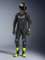 Motorcycle rider wearing the Alpinestars GP Force V2 Leather Suit in Black, 1-piece leather suit with white branding, paired with a black and fluo yellow racing helmet, gloves, and boots, professional track gear on a grey studio background
