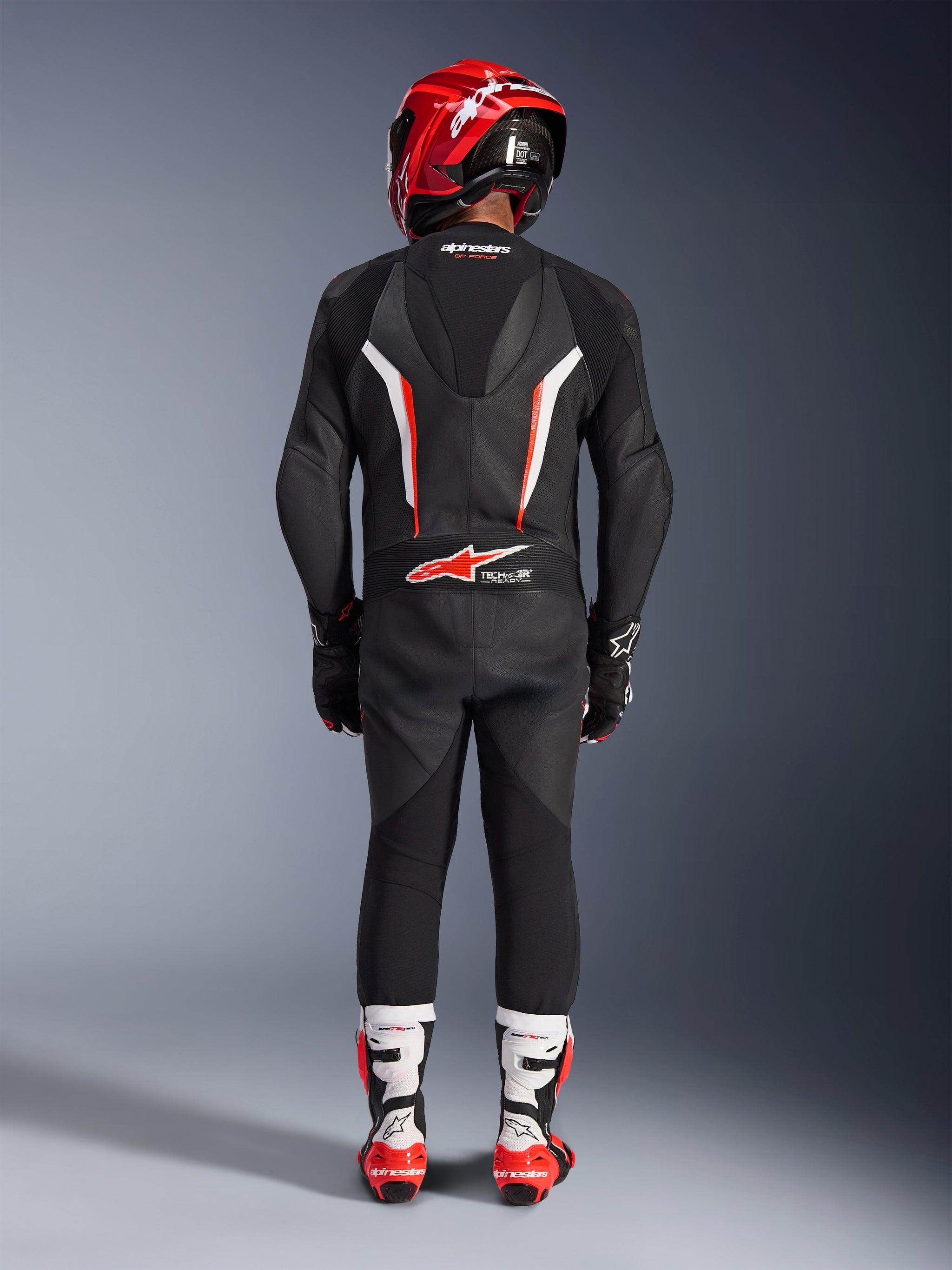 Rider viewed from behind wearing the Alpinestars GP Force V2 Leather Suit 1-Piece in Black White Red Fluo, featuring an aerodynamic speed hump and Tech-Air Ready branding, coordinated with a red racing helmet and white and red boots, professional track gear design.