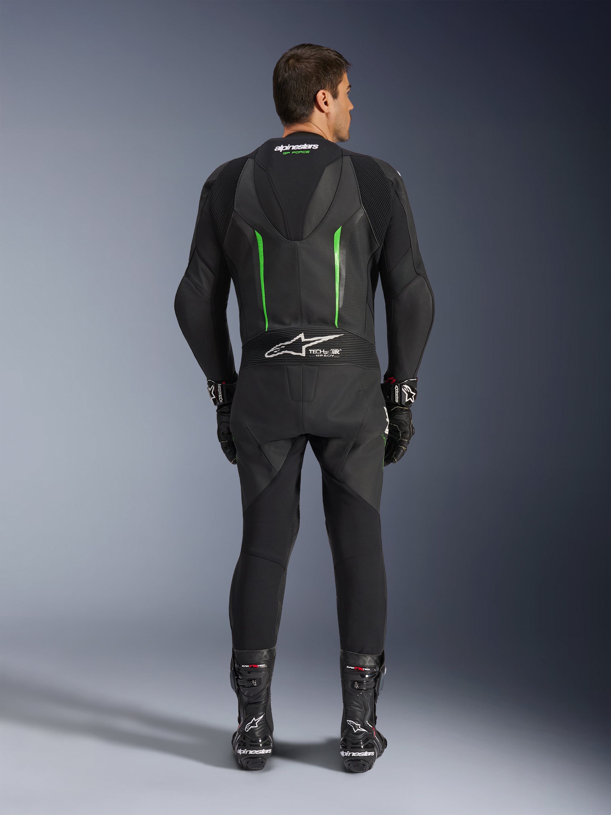 Rear view of the Alpinestars GP Force V2 1-Piece Leather Suit in Black and Green, featuring an aerodynamic speed hump, perforated panels, and Tech-Air Ready branding on the lower back, worn with black racing gloves and boots.