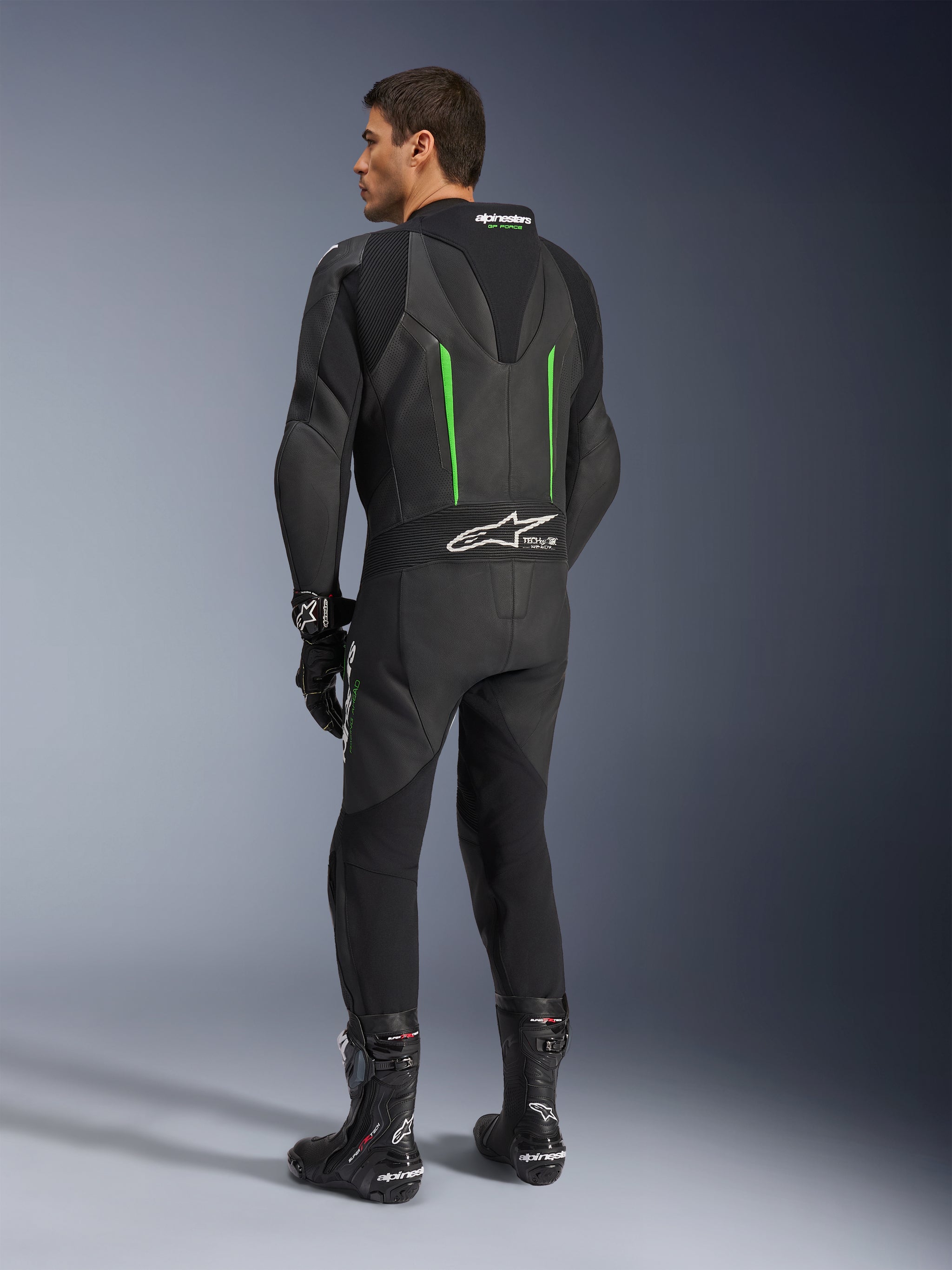 Alpinestars GP Force V2 Leather Suit, 1-Piece Motorcycle Suit, Black with Green accents, back view showing the aerodynamic speed hump and white logo on the accordion stretch panel, paired with black racing boots and gloves.