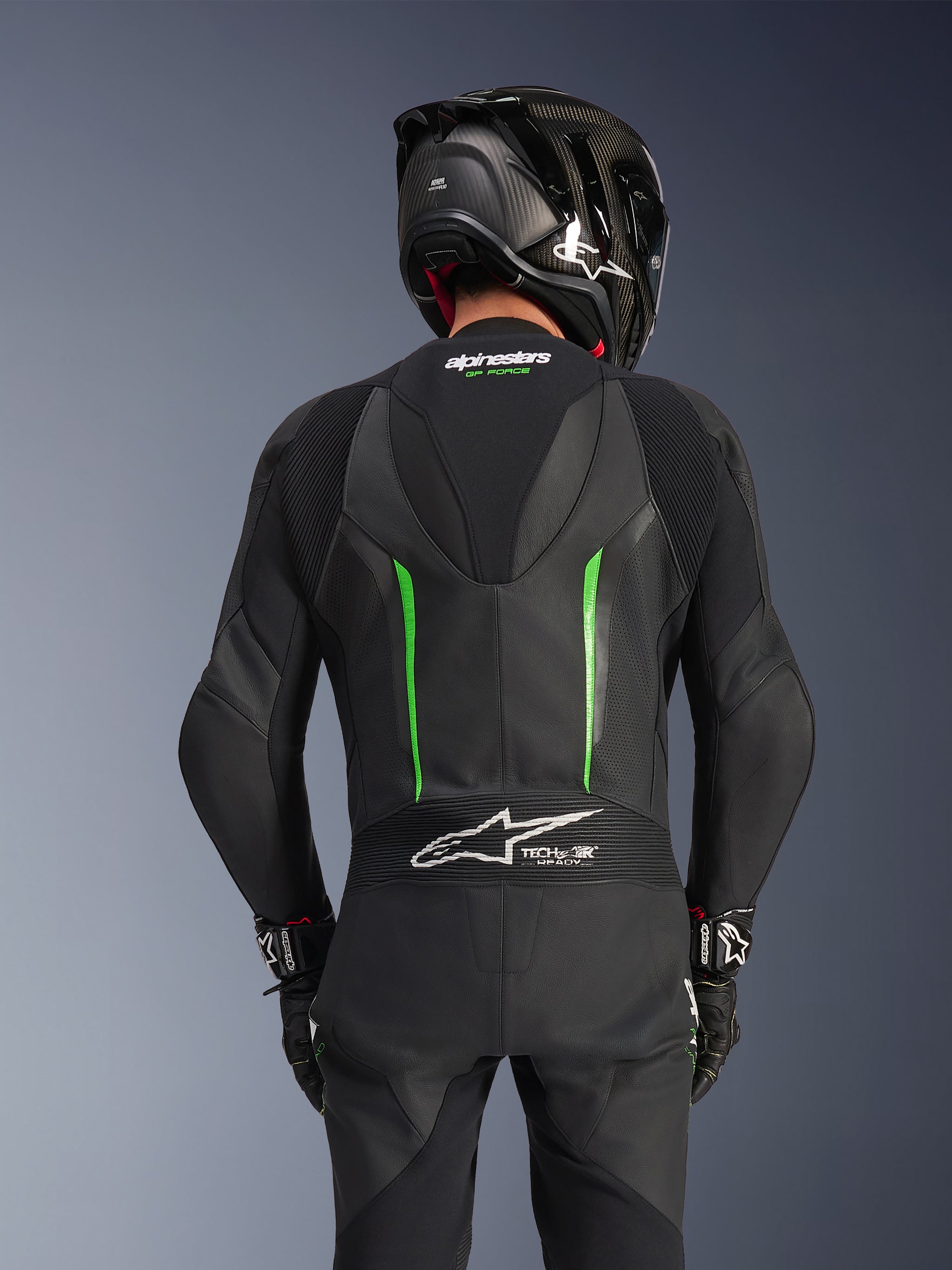 Rear view of a rider wearing the Alpinestars GP Force V2 Leather Suit in Black Green, featuring an aerodynamic speed hump, Tech-Air Ready branding on the lower back accordion panel, and perforated leather construction, paired with a black carbon fiber racing helmet.