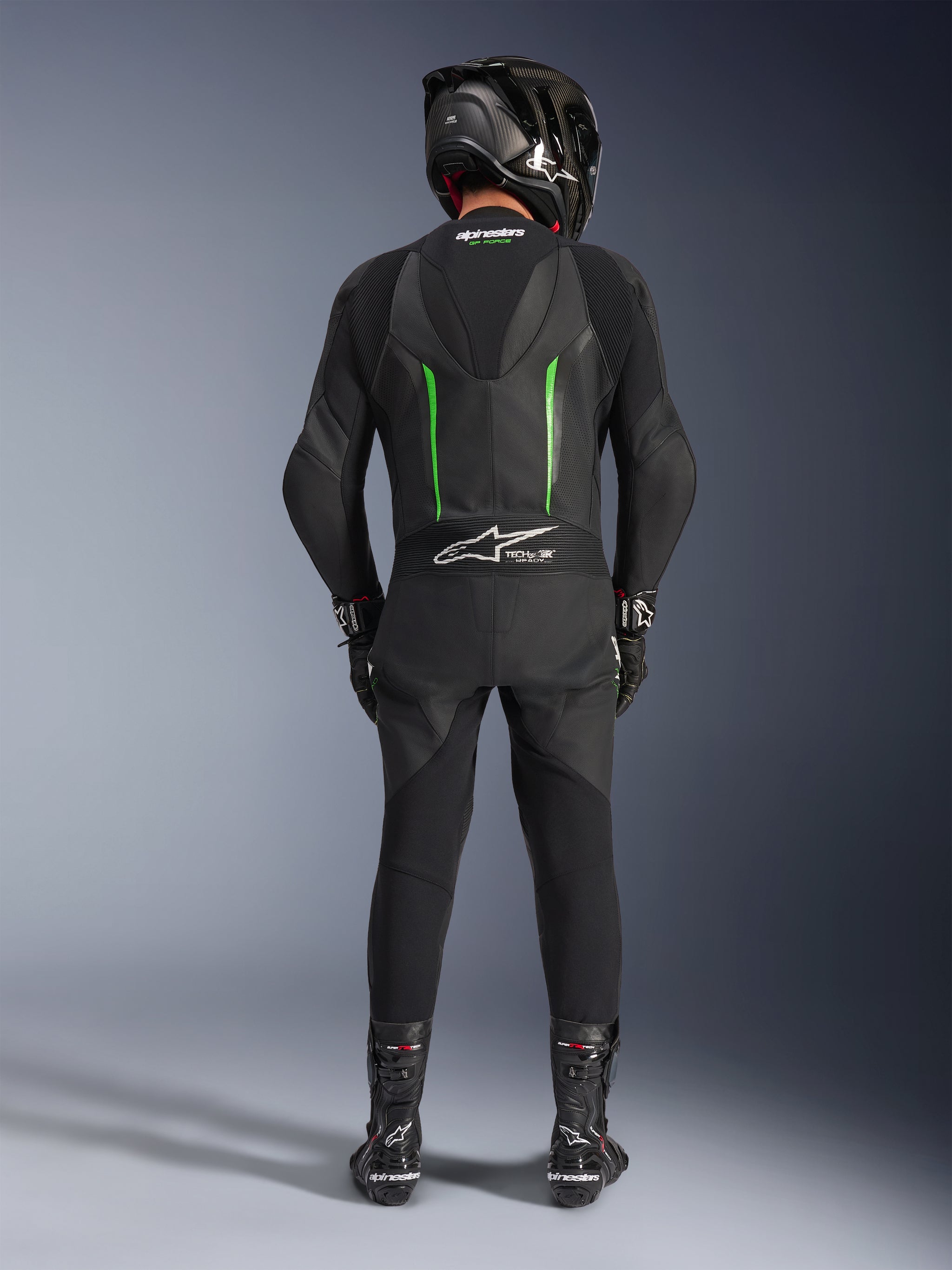 Rider from the back wearing Alpinestars GP Force V2 Leather Suit - 1Piece in Black and Green, paired with a black carbon fiber helmet, black racing gloves, and black boots, featuring an aerodynamic speed hump and Tech-Air Ready branding.