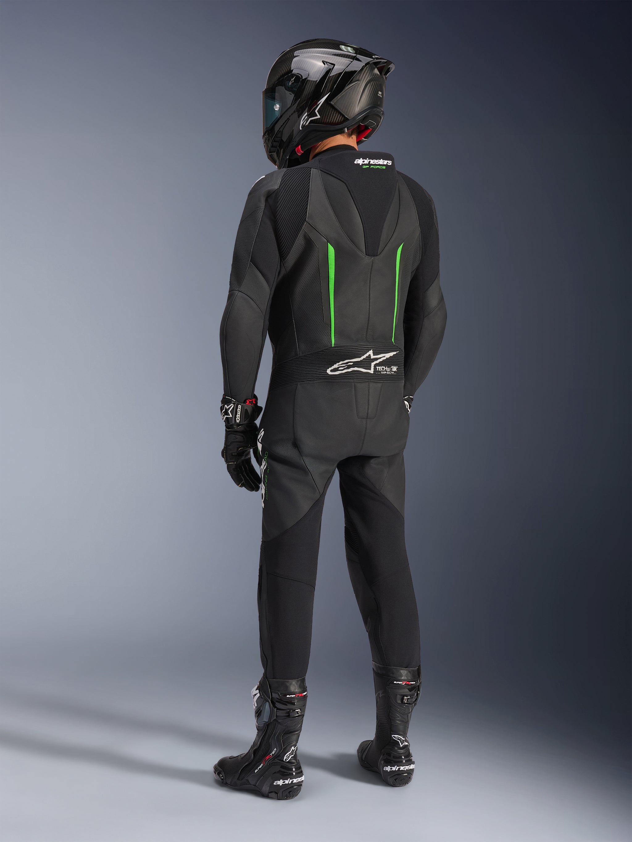 Rider from behind wearing Alpinestars GP Force V2 Leather Suit, 1-Piece, Black with Green accents, aerodynamic speed hump, paired with a black carbon-fiber helmet, black racing boots, and gloves, high-performance motorcycle racing gear