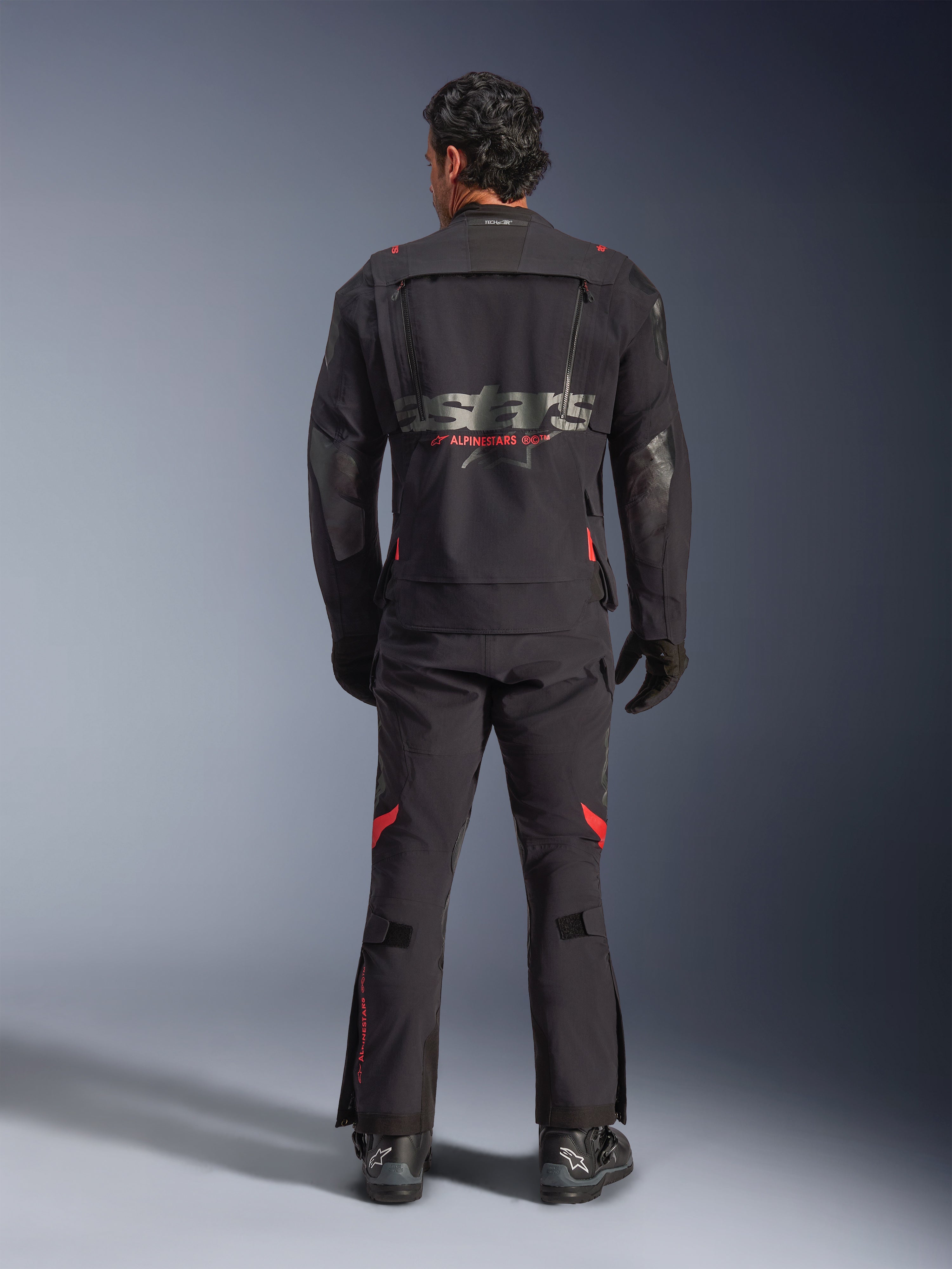 A rider seen from behind wearing the Alpinestars Halo Pro Drystar® XF Laminated Jacket in Black Dark Shadow, featuring a large gray astars logo with red Alpinestars branding, modular ventilation panels on the back, and paired with matching adventure riding pants and black boots.