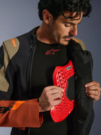 Alpinestars Halo Pro Drystar® XF Laminated Jacket in Walnut Rust Black, lifestyle shot of a male model unzipping the adventure jacket to reveal a red chest protector insert, featuring orange branding on the shoulder and a black base layer with a red logo.