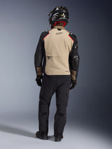 Rider from the back wearing the Alpinestars Andes Pro Drystar® XF Jacket in Walnut Aluminum, tan back panel with black sleeves and red accents, paired with black technical adventure pants and a motorcycle helmet, premium all-weather touring gear.