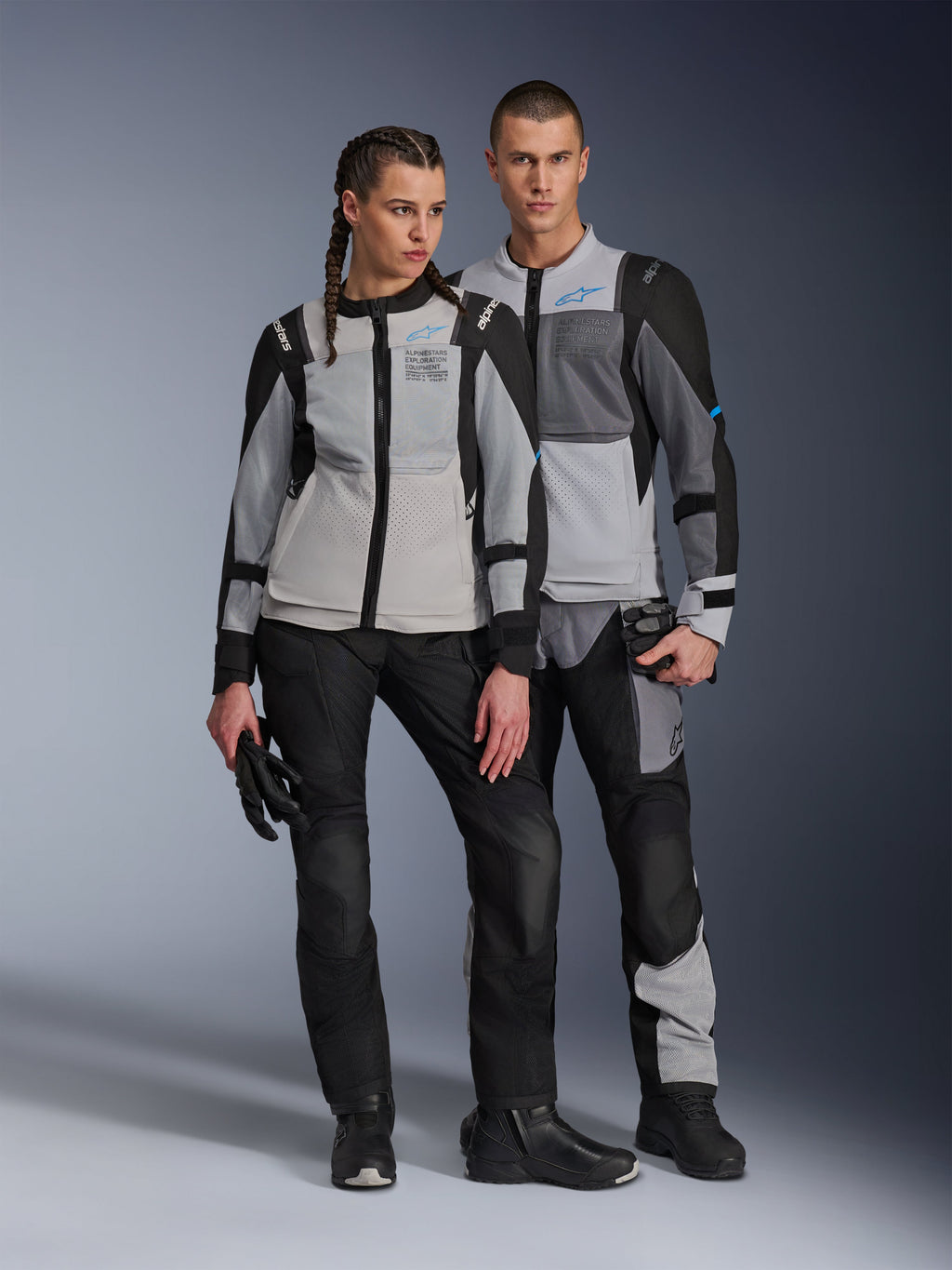 Male and female models wearing Alpinestars ST-2 Air Jackets in Ice Gray Dark Gray and Black, technical summer touring motorcycle gear featuring large mesh panels and exploration equipment branding, paired with black riding pants, gloves, and boots.
