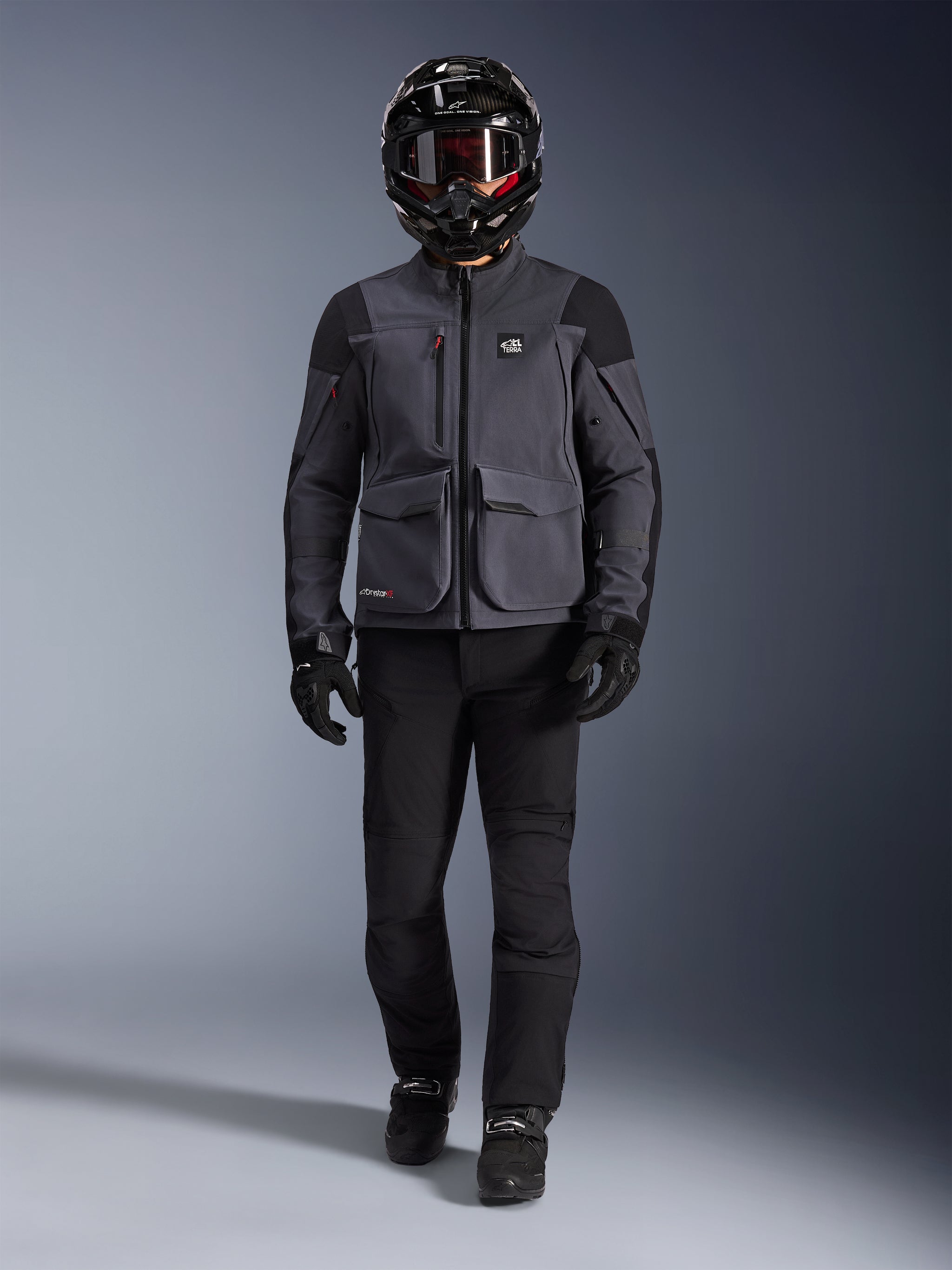 Alpinestars Maxdura Drystar®XF Laminated Armored Shell, adventure motorcycle jacket in Deep Gray and Black, worn with matching black riding pants, a black full-face helmet, gloves, and boots, featuring prominent cargo pockets and ALL TERRA branding for a technical off-road touring look