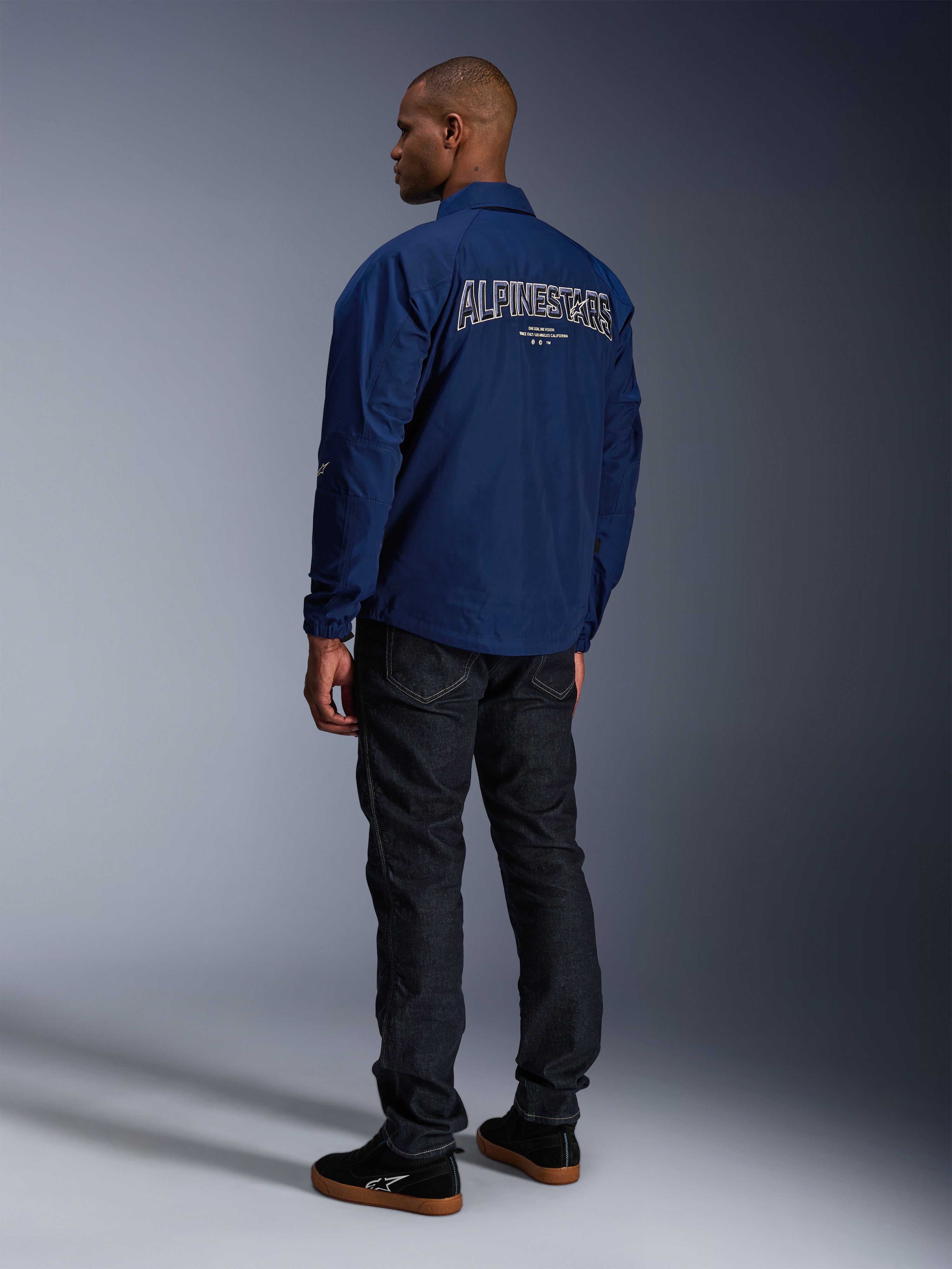 Model wearing the Alpinestars Revved WR Jacket in Navy Blue from behind, featuring large white outlined 'ALPINESTARS' embroidery across the back, paired with dark denim pants and black sneakers, urban motorcycle aesthetic.