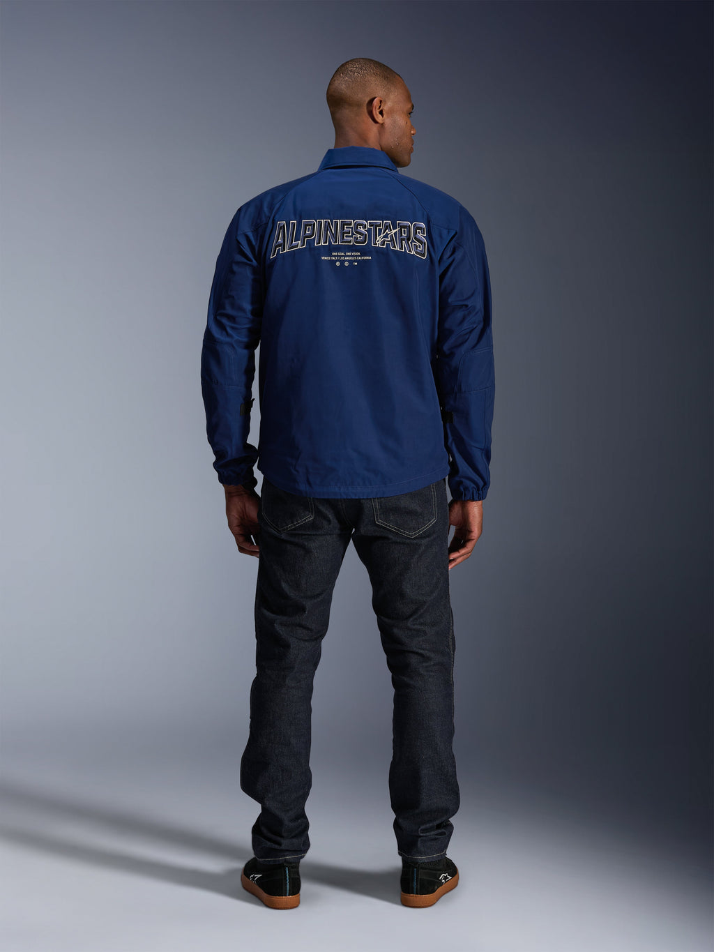 Alpinestars Revved WR Jacket in Navy Blue, rear view, featuring large 3D effect 'ALPINESTARS' embroidery across the back, styled with dark denim jeans and black sneakers, urban motorcycle apparel.
