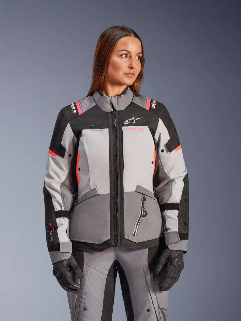 Alpinestars Stella Andes V4 Drystar® Jacket, Women's Motorcycle Jacket, Dark Gray Ice Gray Black Coral Fluo, worn by a female model with matching adventure pants and black gloves, featuring a multi-tonal gray textile design with vibrant coral accents, designed for all-weather adventure touring.