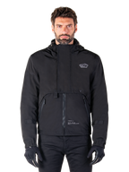 Alpinestars Mootant Waterproof Parka, Black hooded motorcycle jacket with a modular technical design, worn by a male model with black gloves, featuring a unique chest flap and zippered side pockets, urban and touring riding style.