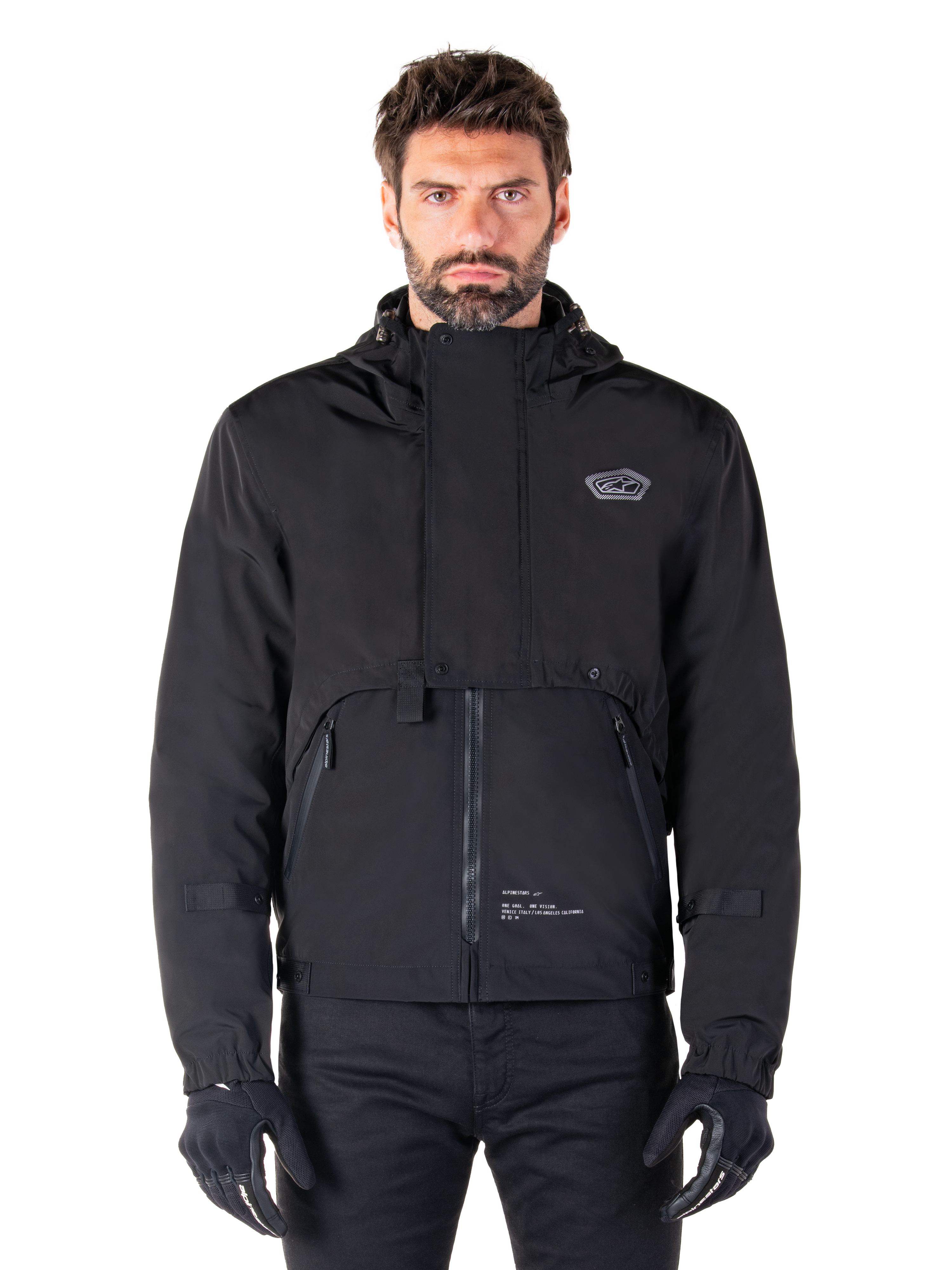 Alpinestars Mootant Waterproof Parka, Black hooded motorcycle jacket with a modular technical design, worn by a male model with black gloves, featuring a unique chest flap and zippered side pockets, urban and touring riding style.