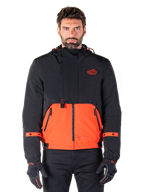 Alpinestars Mootant Waterproof Parka, modular motorcycle jacket in Spicy Orange and Black, worn by a model with matching gloves, features a hooded design with two-tone panels, front zipper, and adjustable cuffs, urban contemporary riding apparel
