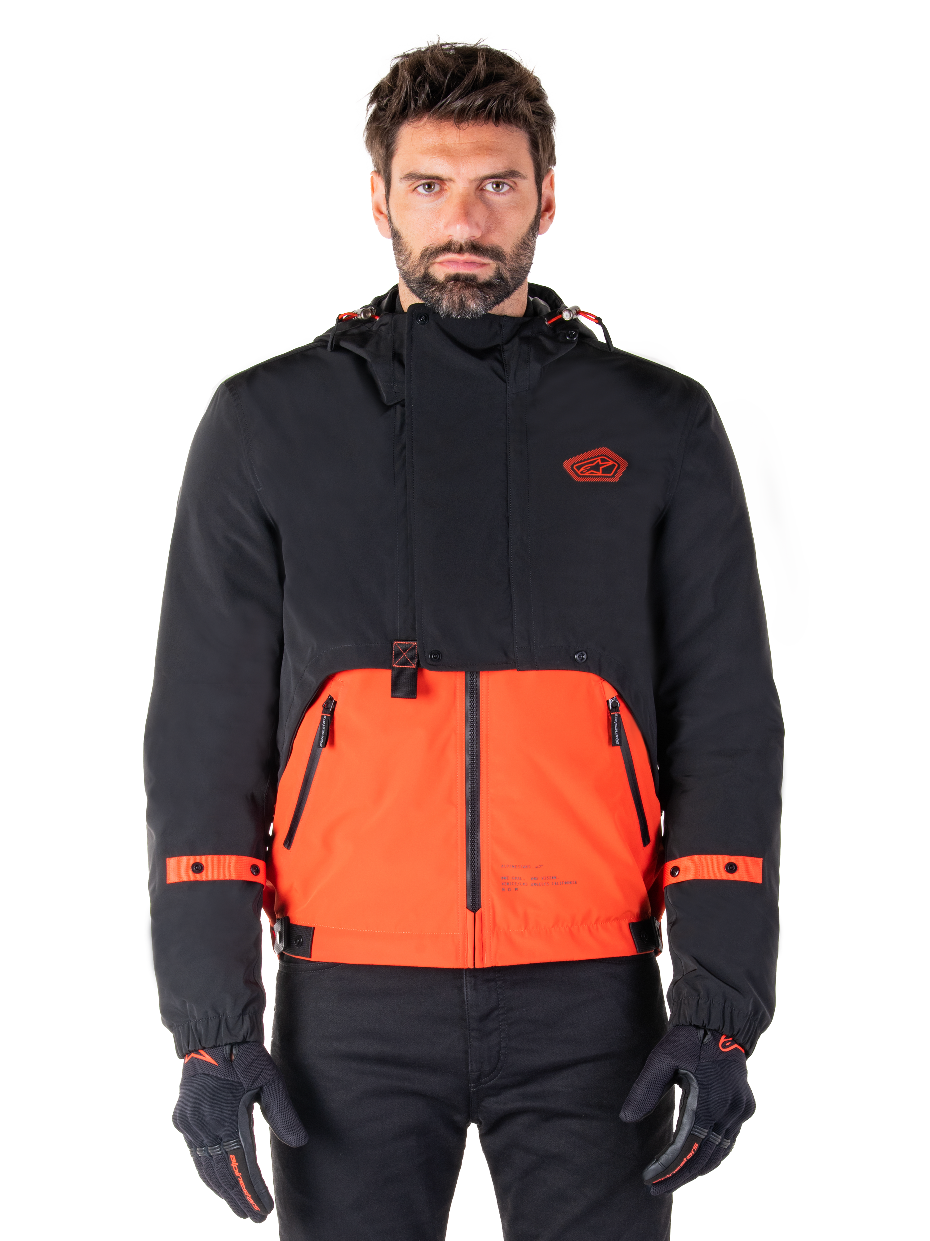 Alpinestars Mootant Waterproof Parka, modular motorcycle jacket in Spicy Orange and Black, worn by a model with matching gloves, features a hooded design with two-tone panels, front zipper, and adjustable cuffs, urban contemporary riding apparel