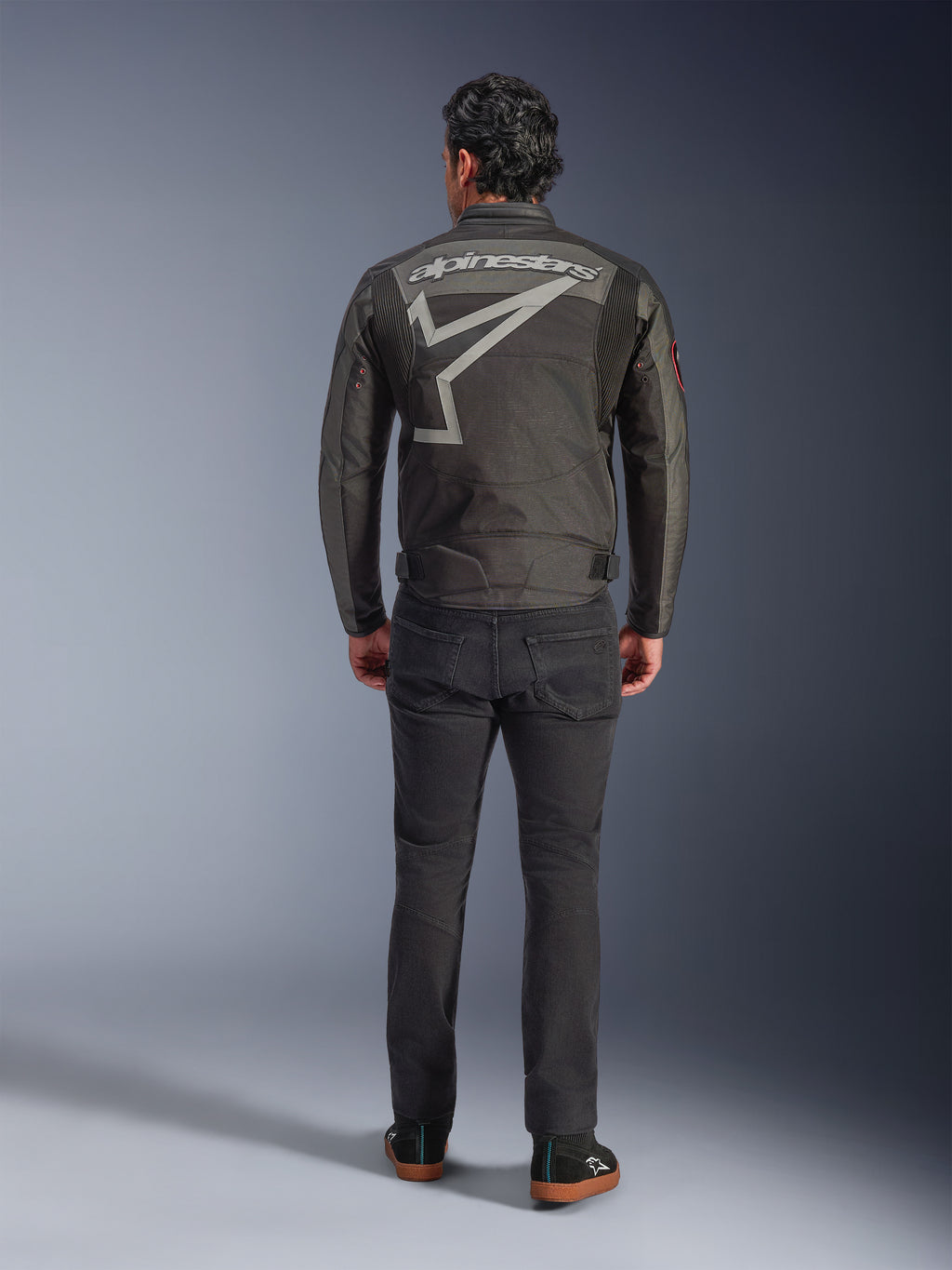 Alpinestars Axton Jacket, Motorcycle Jacket, Black Anthracite Red Tangerine, back view showing large grey Alpinestars logo and star graphic, accordion shoulder panels, worn with black pants and casual black shoes.