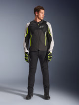 Male model wearing Alpinestars AST V3 Air Jacket in Black Silver Yellow Fluo, featuring mesh ventilation panels and fluorescent accents, paired with black riding pants and matching motorcycle gloves, sport-riding performance gear.