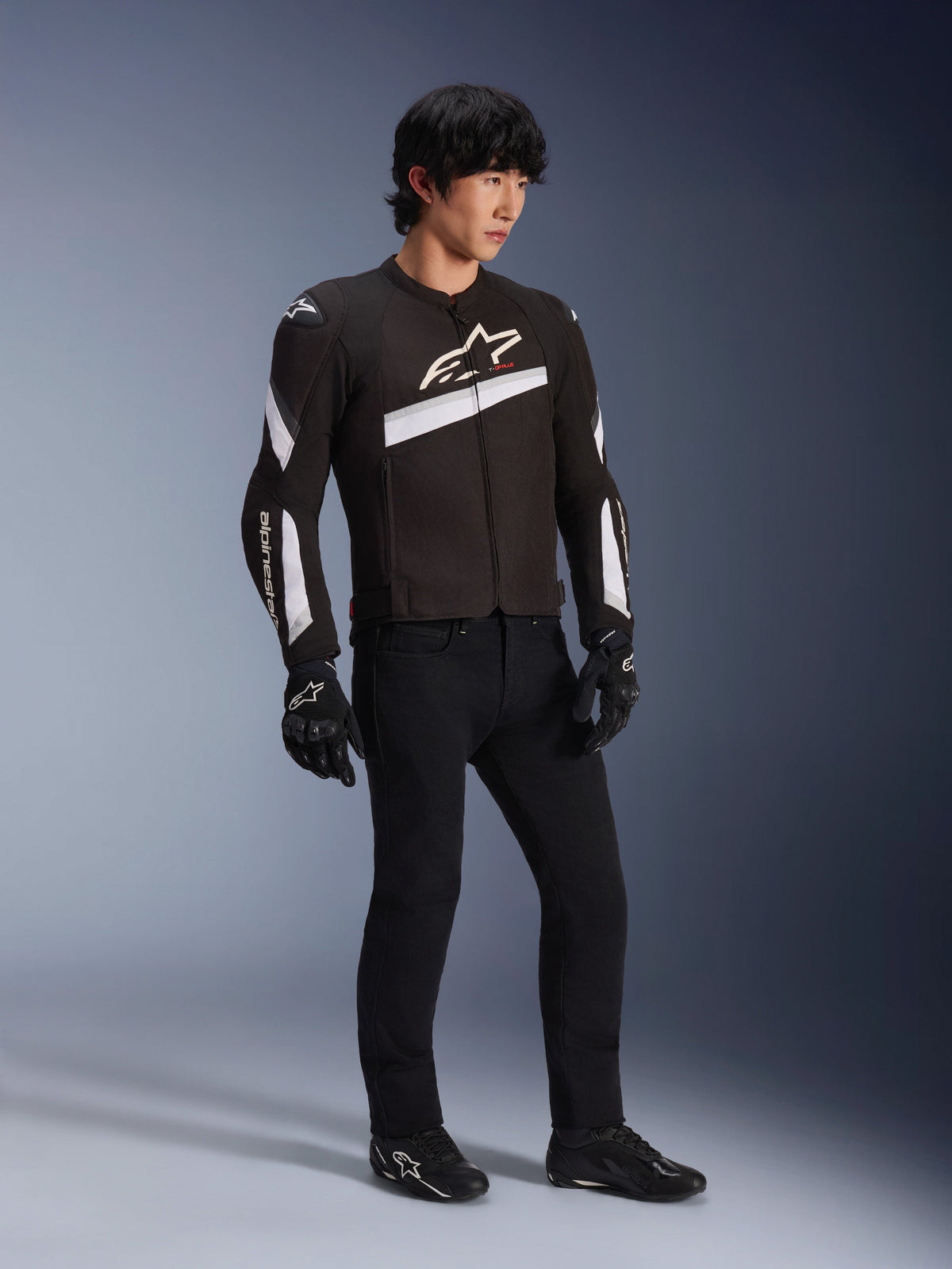 Alpinestars T-GP Plus R V4 Jacket, Black and White textile motorcycle jacket worn by a model with black riding pants and gloves, featuring white Alpinestars logos on the chest and sleeves, sporty design for road performance