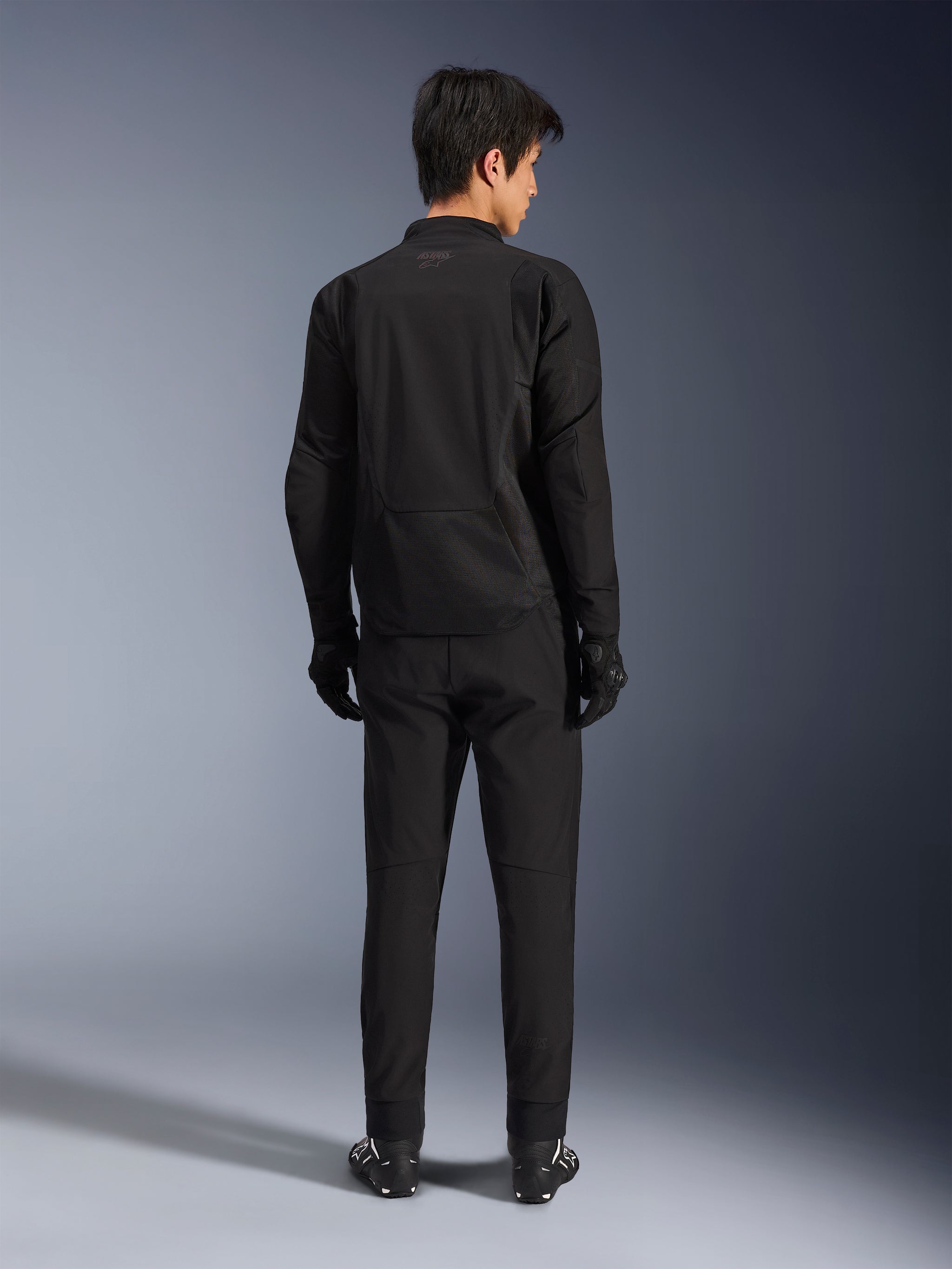 Alpinestars Aeroshell Airflow Jacket in Black Anthracite, back view of a male model wearing full motorcycle riding gear including black pants and gloves, featuring a lightweight softshell construction with an elongated rear for coverage.