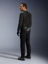 A man wearing the Alpinestars T-SPS Air V2 Jacket in Black Fluo Green, shown from a back and side profile view, styled with black riding pants and motorcycle shoes. The jacket features ventilated tech mesh panels, bright green accents on the shoulders and lower back, and a white star logo on the rear tail.