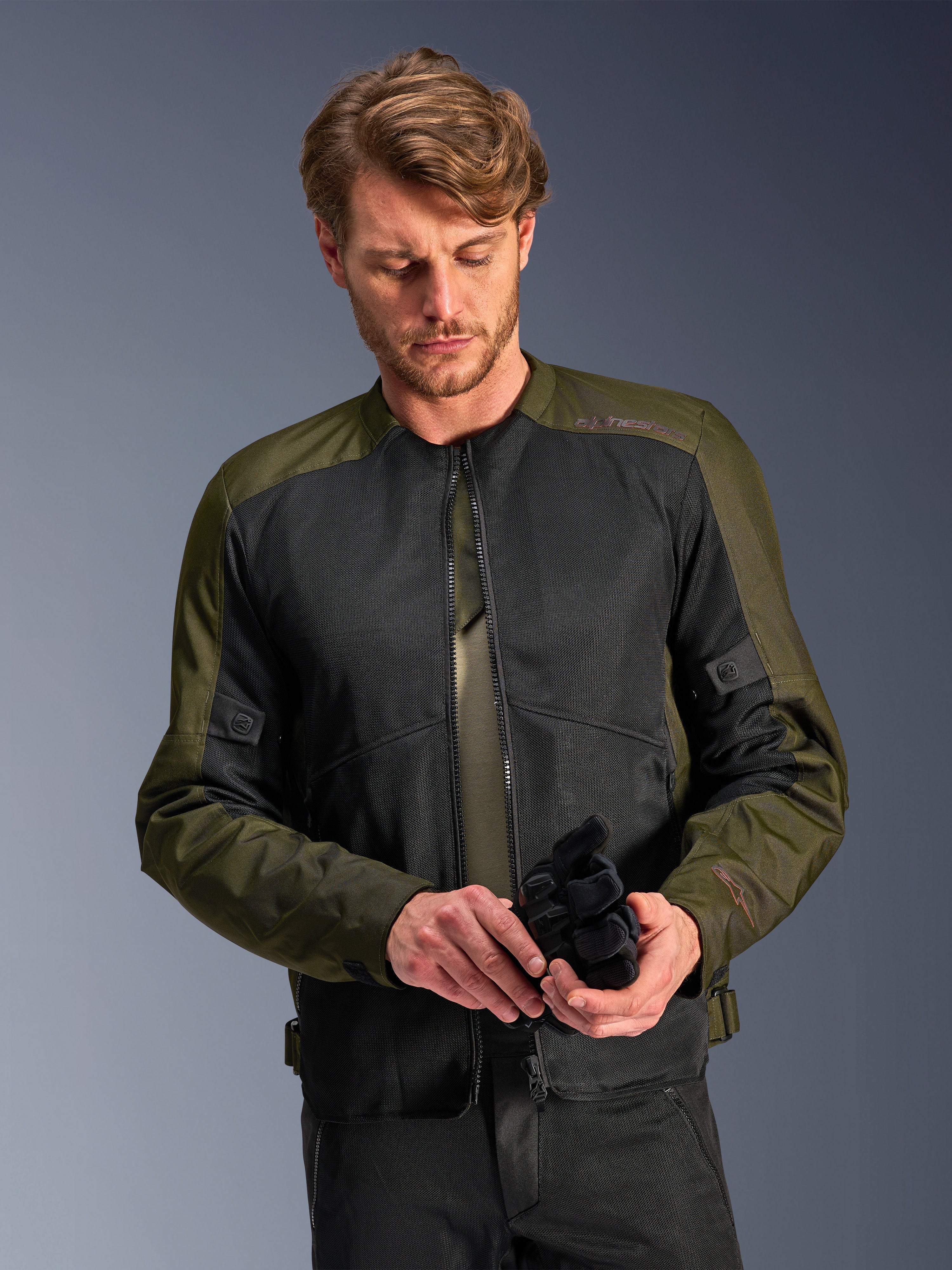 Man wearing the Alpinestars C-1 Air Jacket in Khaki, urban motorcycle jacket with olive green textile and black mesh panels, looking down while holding black motorcycle gloves, clean and contemporary style for road riding.