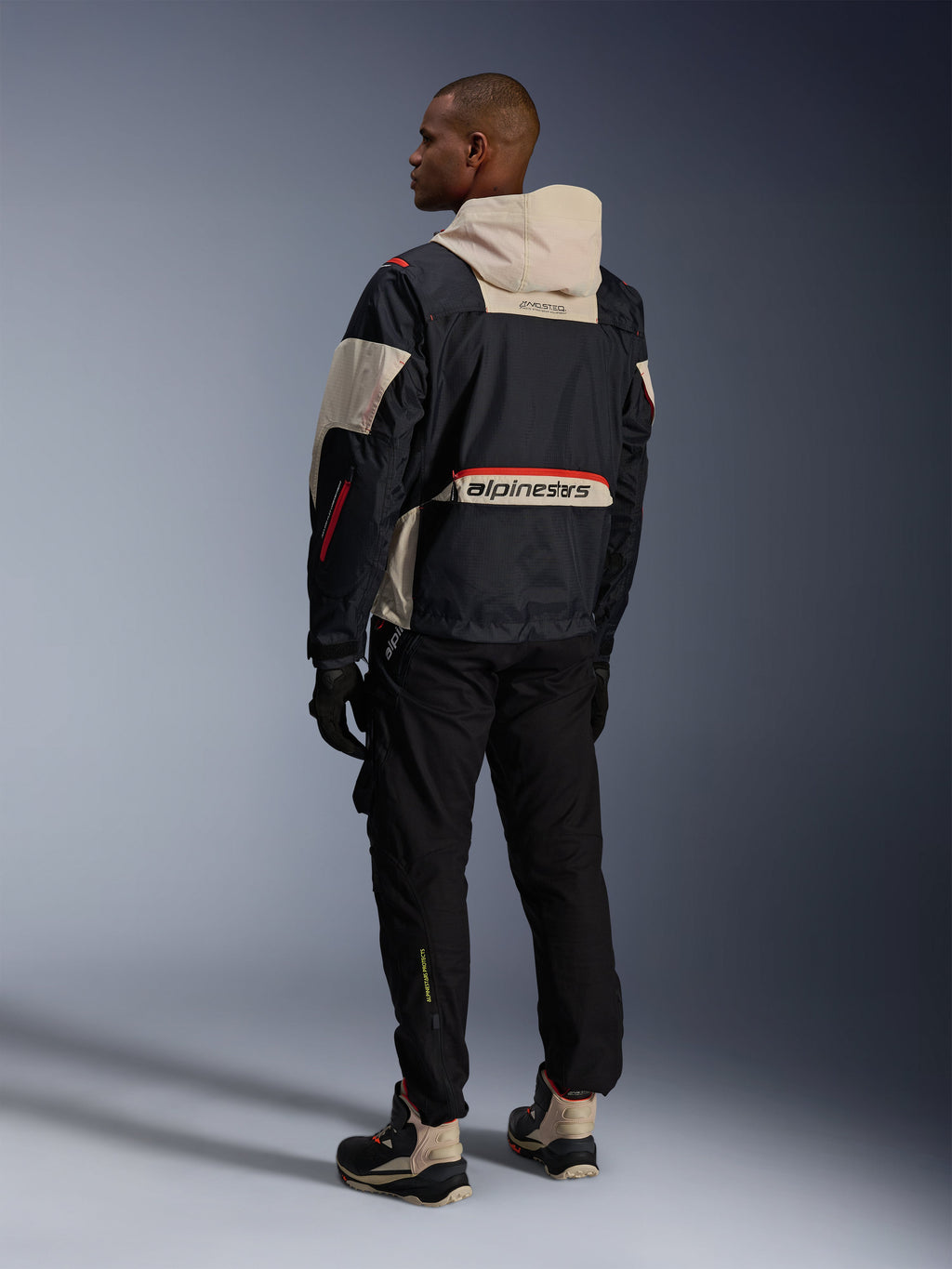 Rear view of a man wearing Alpinestars Acteon Pants in Black, paired with a Mogress Air Jacket in black and cream, black gloves, and riding boots, contemporary urban motorcycle outfit