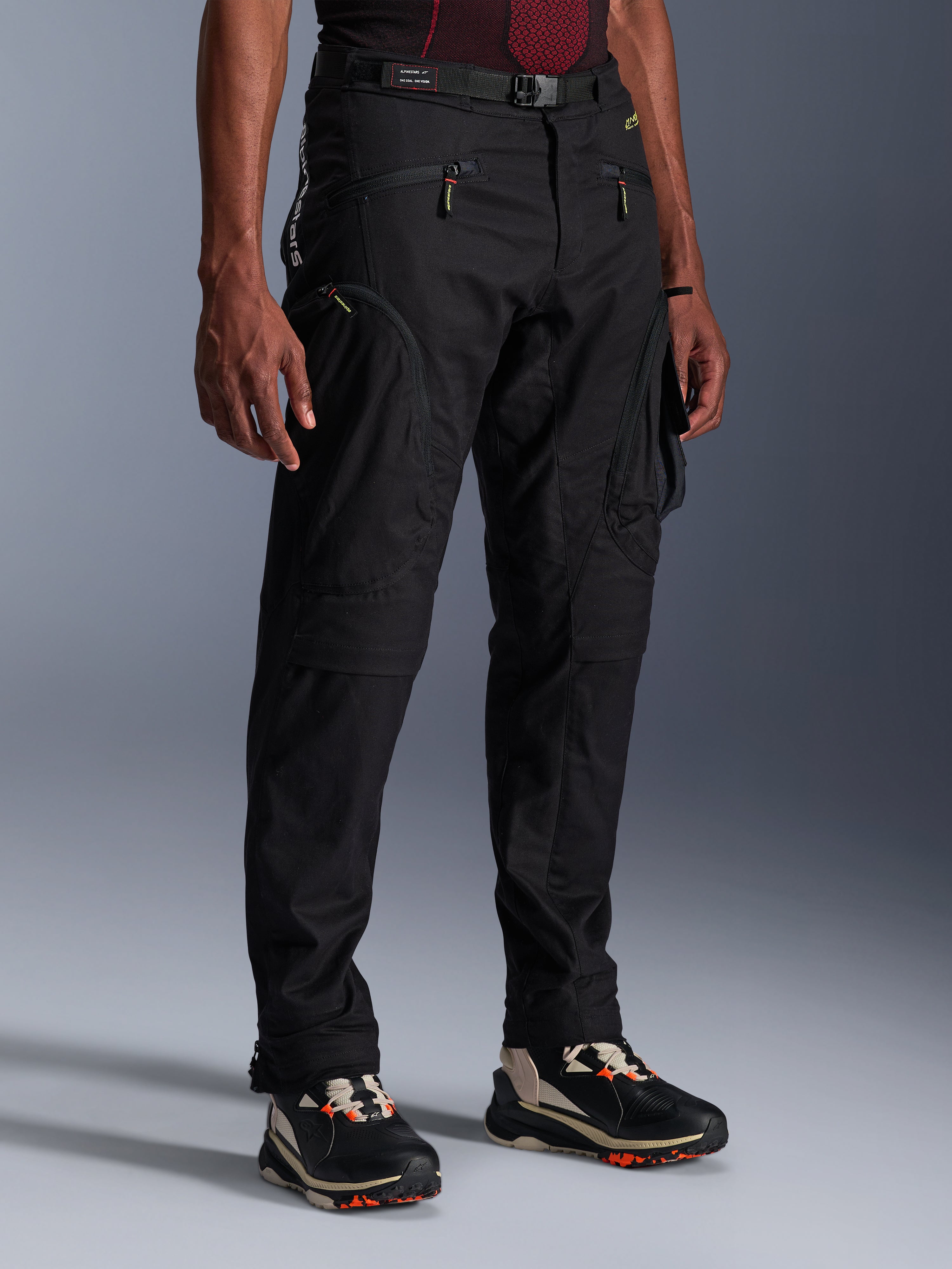Alpinestars Acteon Pants, Black Motorcycle Pants, worn by a model, featuring a modern tapered silhouette, zippered cargo pockets, adjustable waist belt, and technical sneakers, urban riding apparel