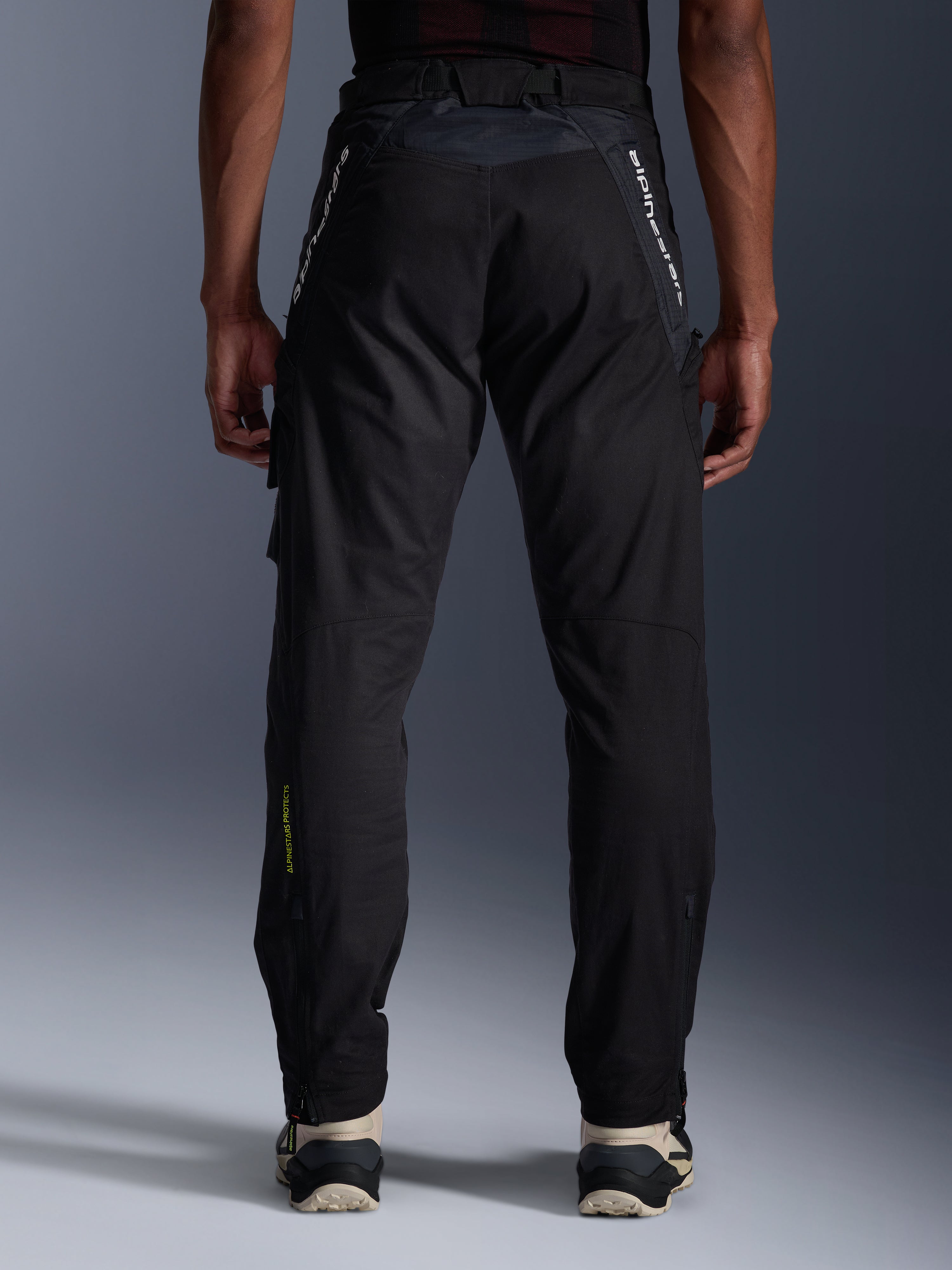 Alpinestars Acteon Pants, Urban Motorcycle Pants, Black, rear view showing tapered fit, white branding on upper legs and yellow logo on the lower calf, adjustable leg openings with zippers, worn with cream sneakers