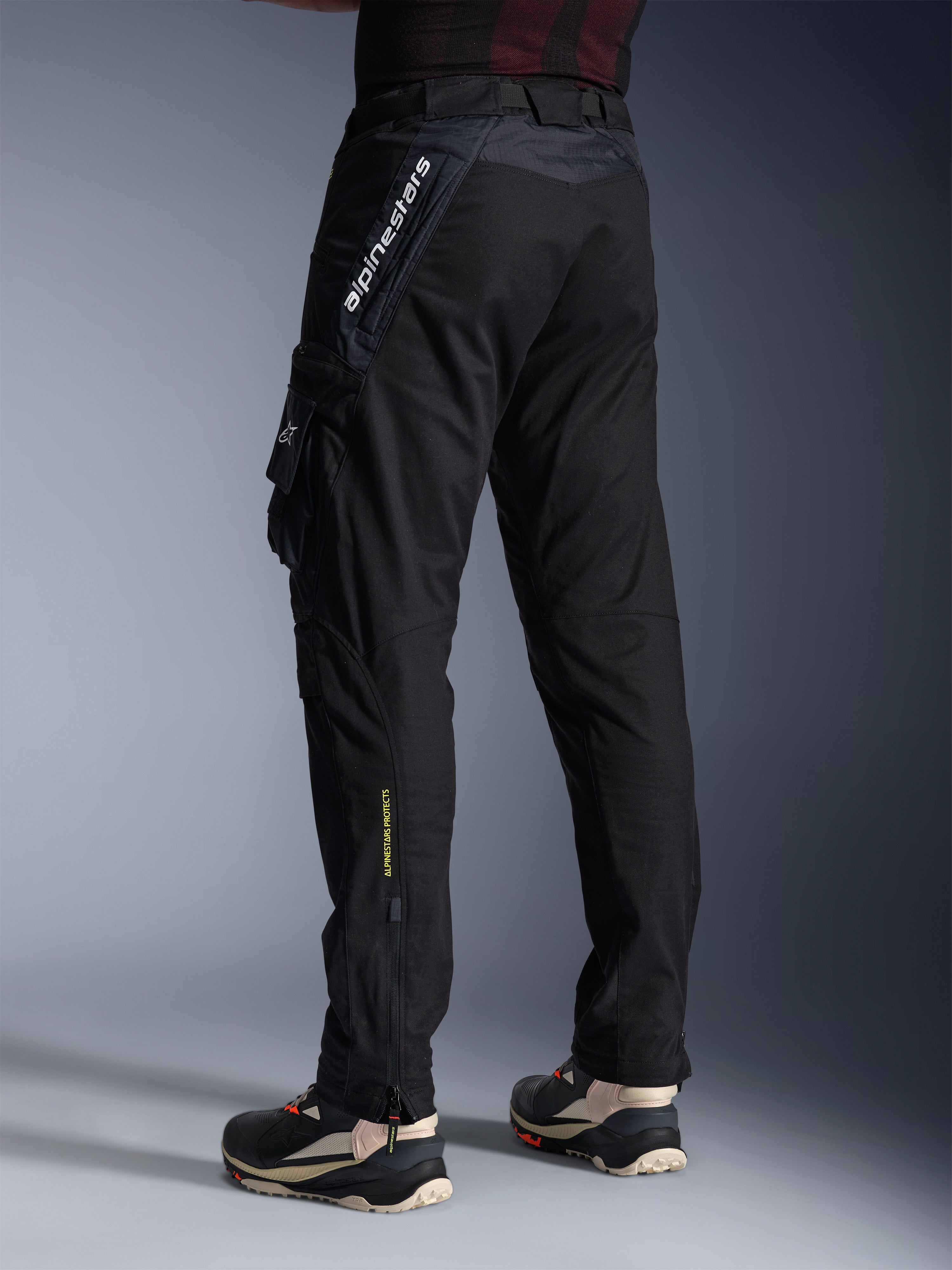 Alpinestars Acteon Pants, Black motorcycle pants, back and side view showing a modern tapered fit, side cargo pocket with logo, vertical white Alpinestars branding on the upper thigh, yellow 'Alpinestars Protects' text on the lower leg, urban contemporary riding gear