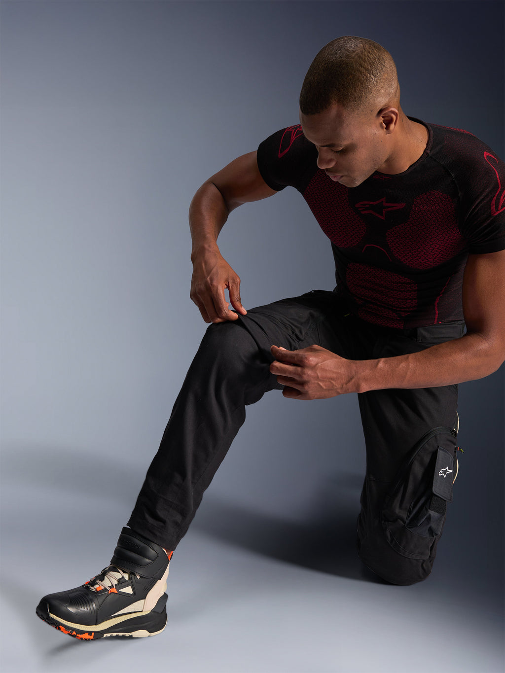 Alpinestars Acteon Pants, Black urban motorcycle pants, worn by a kneeling man adjusting the knee area, featuring cargo pockets and a tapered fit, paired with a black and red Alpinestars base layer and riding shoes