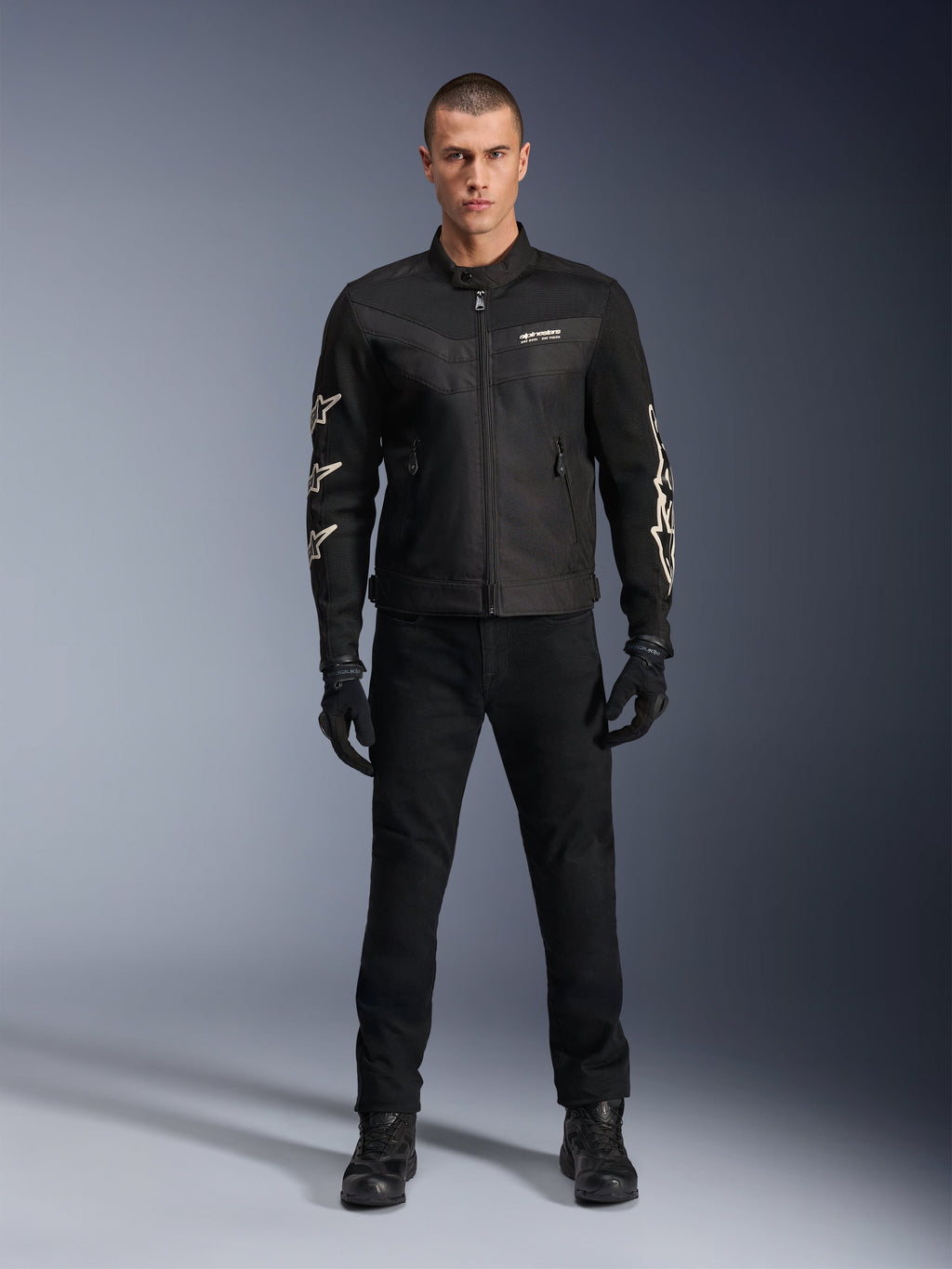 Male model wearing Alpinestars T-Dyno Air Jacket, Black, lightweight textile motorcycle jacket with breathable 3D mesh, white star logos on sleeves, paired with black pants, gloves, and boots, front view standing
