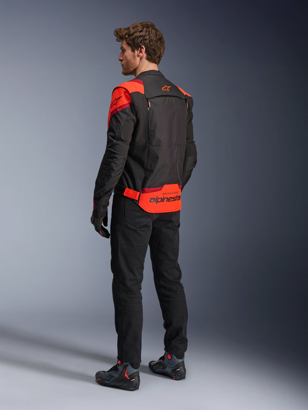 Man wearing Alpinestars T-Stunt Air Jacket from the back, Black Red Fluo and Dark Red, textile motorcycle jacket with mesh panels and sport-cut design, featuring bold Alpinestars branding on the lower back, styled with black pants and riding boots